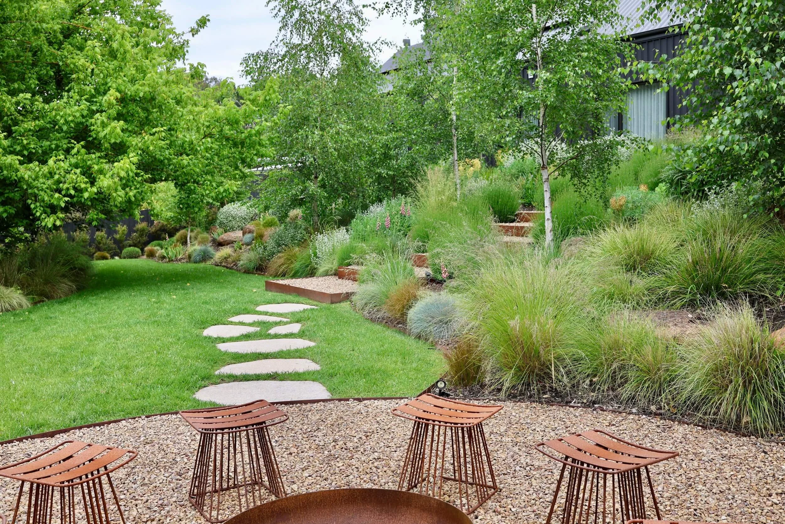 A backyard garden with a stone pathway, surrounded by lush green grass and various plants, trees, and shrubs. There is a patio area with metal chairs and a fire pit in the foreground.