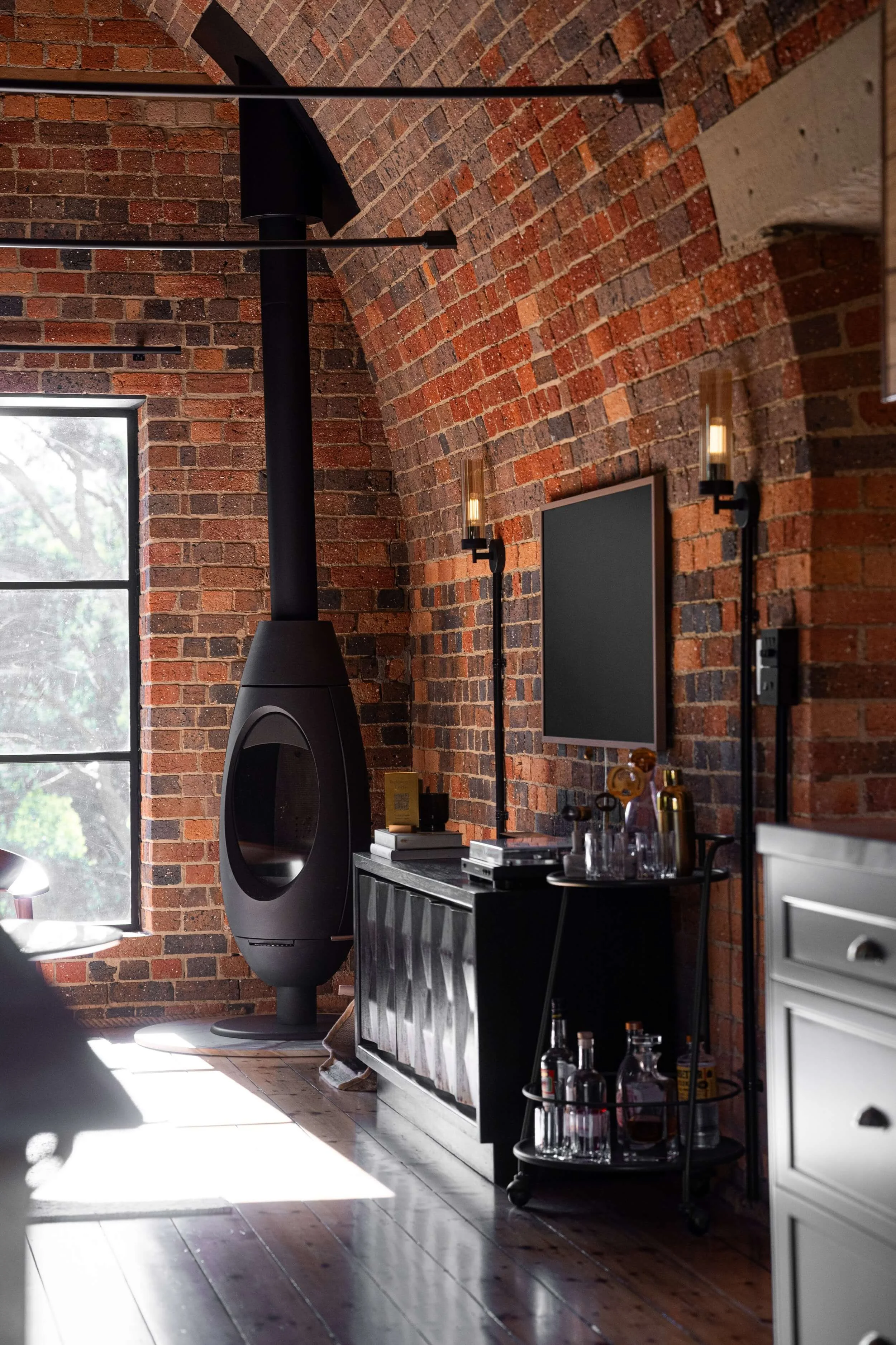 Interior view of a room with brick walls, featuring a black modern fireplace, a window letting in daylight, and a bar cart with glass bottles and glassware. There is a black wall-mounted television and wall sconces with warm lighting