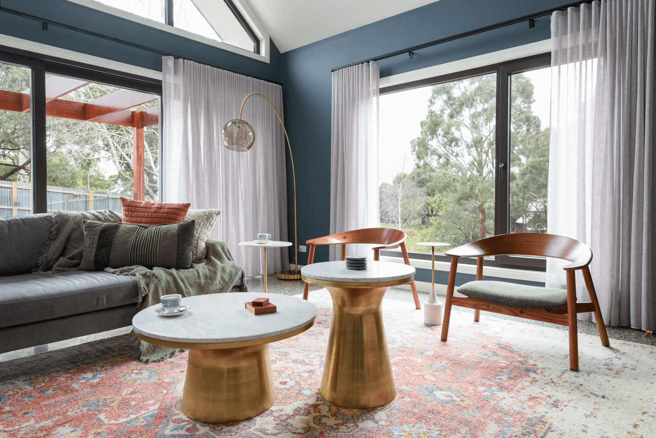 Living room with large windows, blue walls, and white curtains. Contains a gray sofa with pillows and a blanket, two wooden chairs, two small white marble-topped tables with gold bases, a modern arc floor lamp, and a colorful patterned rug. Outside v