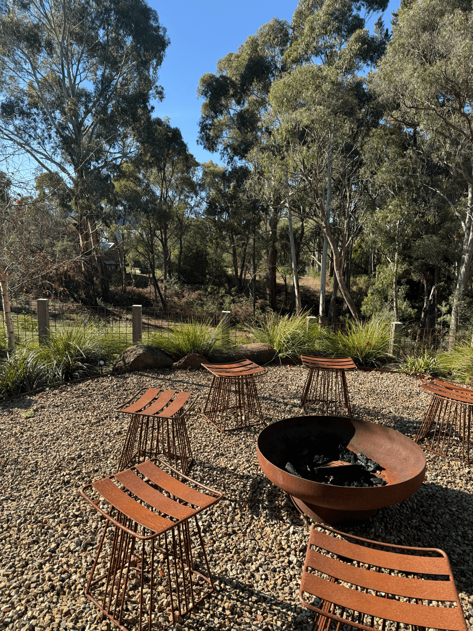 Outdoor seating area with metal chairs around a fire pit on a gravel surface, surrounded by trees and plants under a clear blue sky.