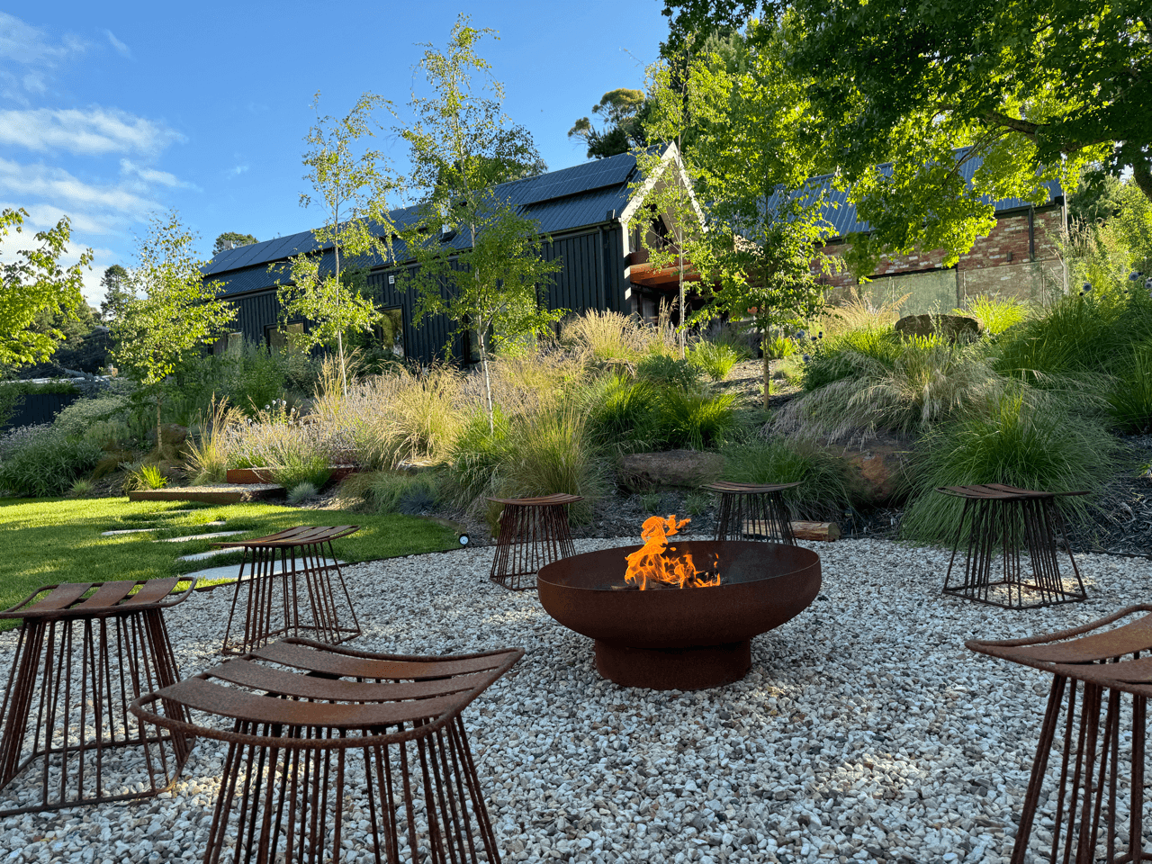 Outdoor garden seating area with a fire pit in the center, surrounded by metal stools on a gravel surface, with lush greenery, trees, and a dark building in the background under a clear blue sky.