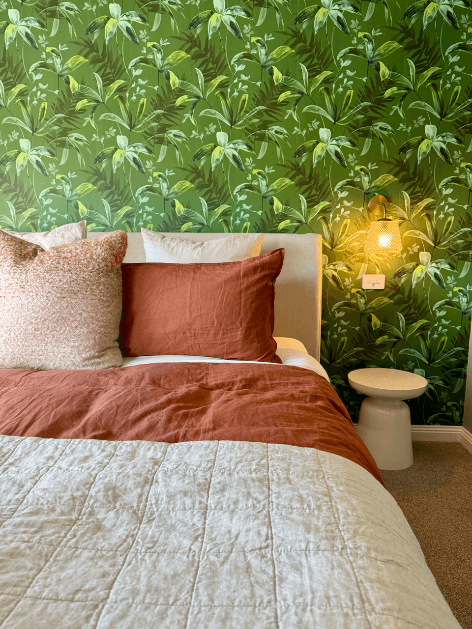 A bedroom with a botanical green leafy wallpaper, a bed with rust-colored and beige pillows, a beige quilted bedspread, a small white side table, and a wall-mounted yellow lamp.