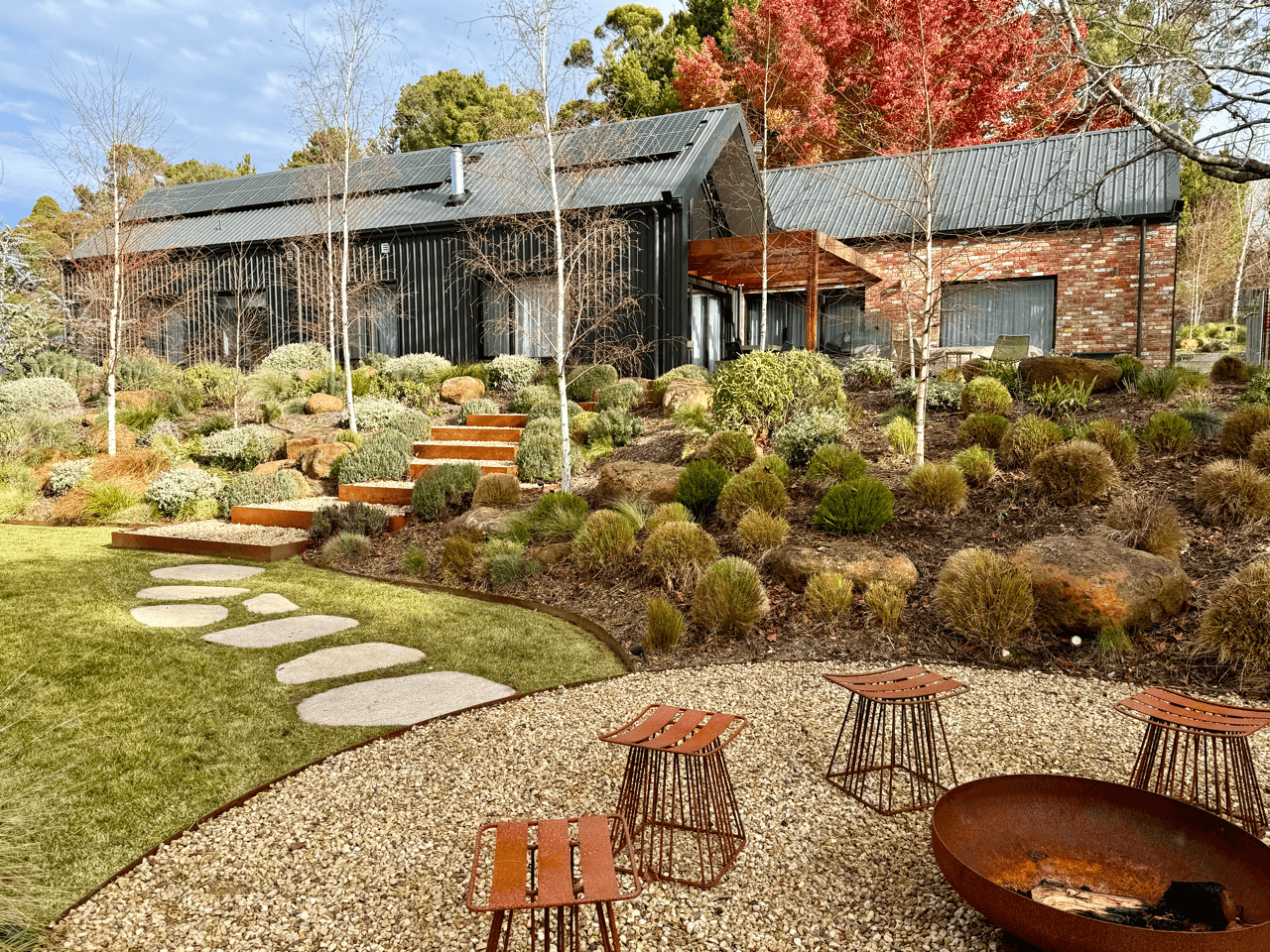 A backyard garden with stone pathways, desert plants, and a modern house with black metal siding and brick walls. There is outdoor seating and a fire pit in the foreground.