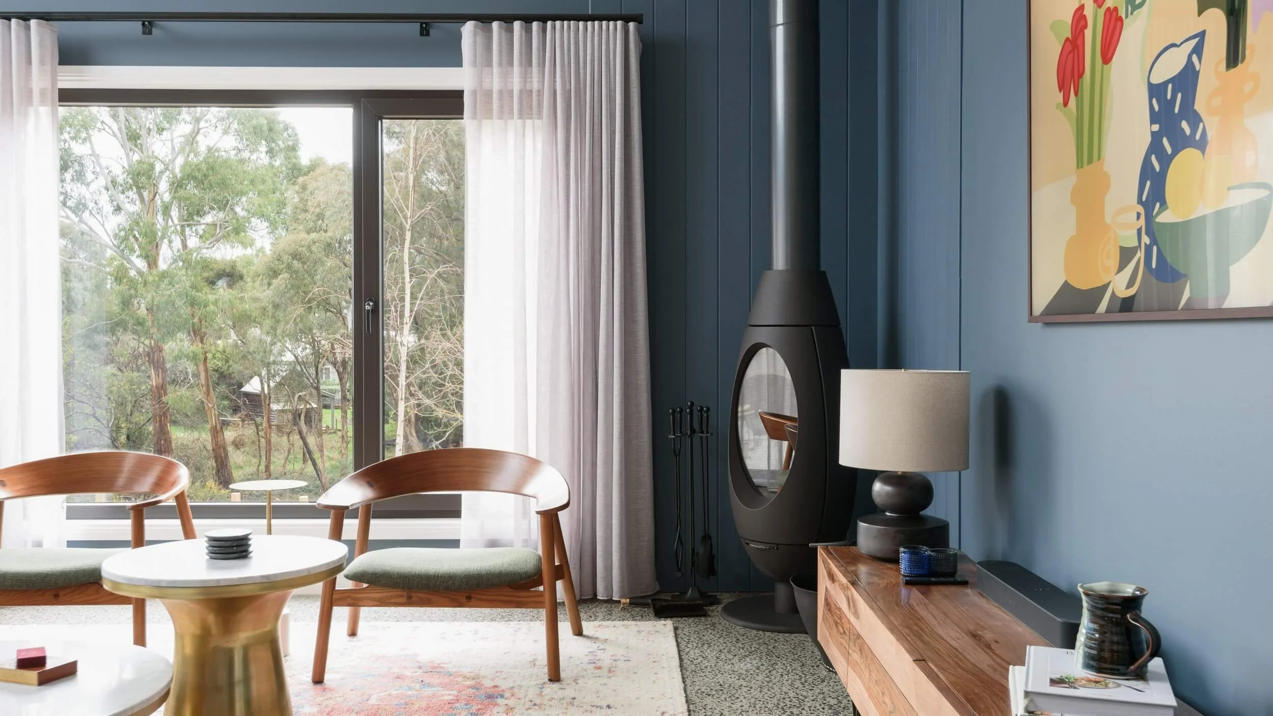 A modern living room with a large window showing trees outside, blue walls, a wooden sideboard, a black fireplace, a white lamp, and colorful art on the wall.