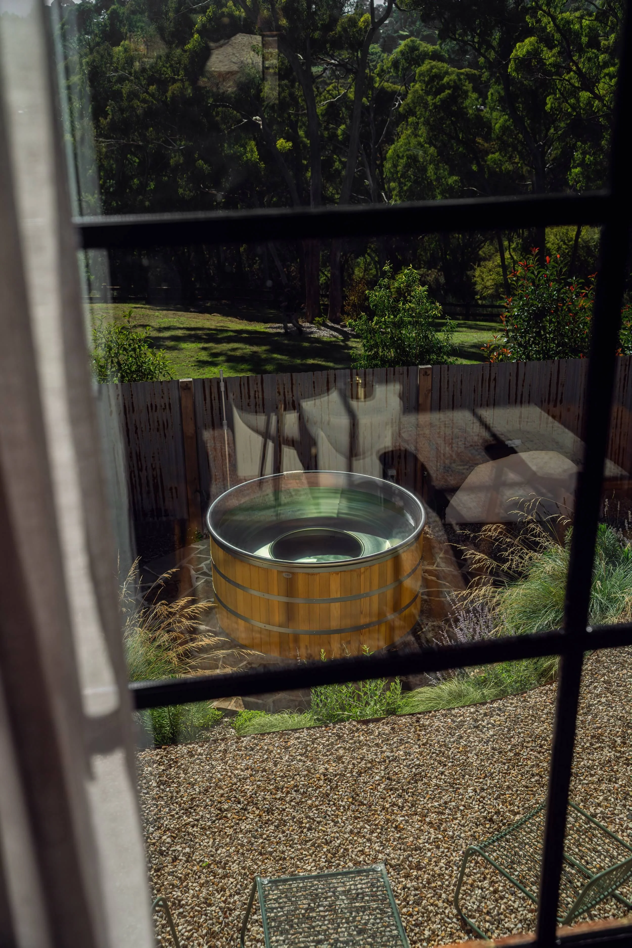 View of a backyard garden through a window with black bars, featuring a hot tub, lawn, trees, and outdoor furniture.