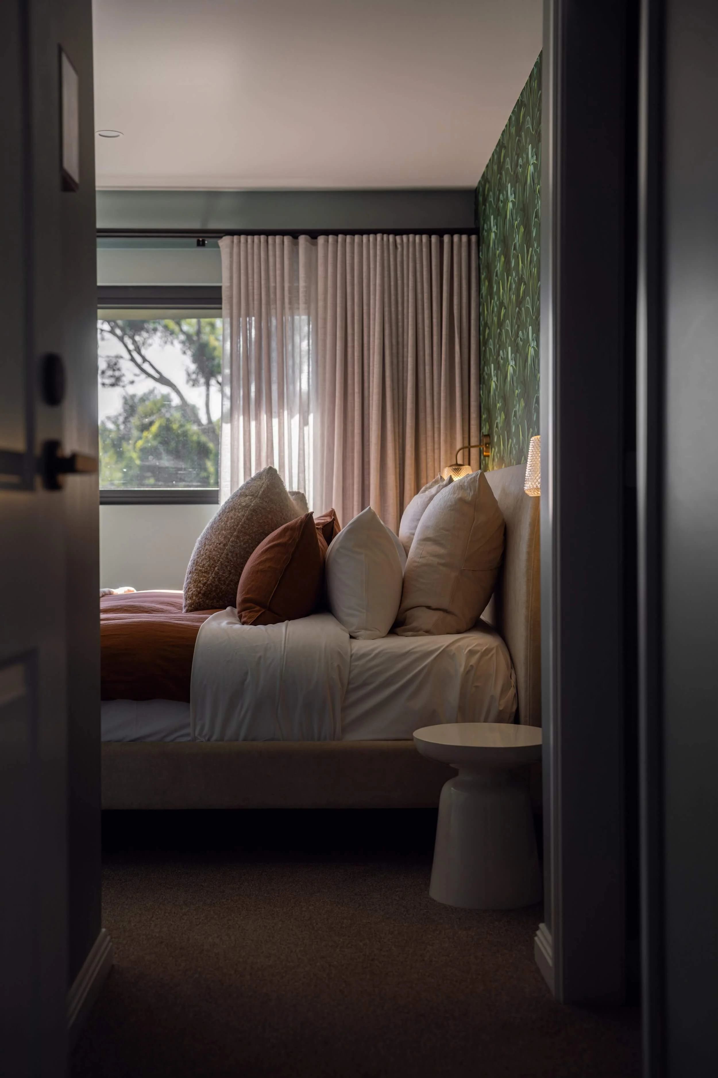 View of a stylish bedroom with a bed adorned with multiple pillows, a bedside table, a large window with beige curtains, and a green leafy wallpaper accent wall, seen through an open doorway.