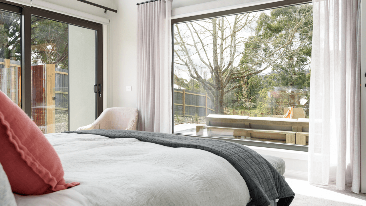 A bedroom featuring a large bed with white bedding and a red pillow, near a sliding glass door leading to an outdoor patio with a wooden bench and outdoor furniture, and large windows with white curtains revealing a garden with trees.