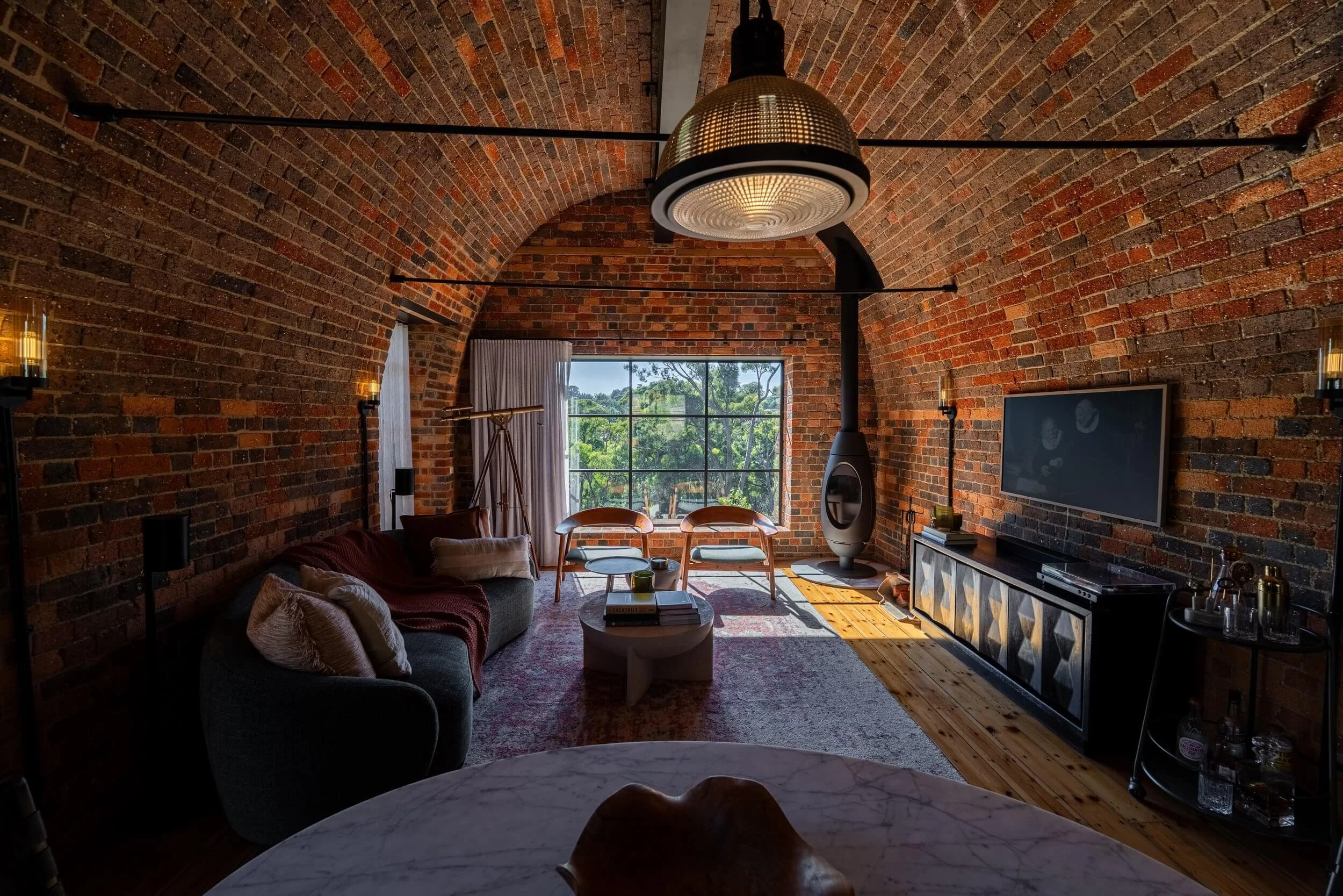 A cozy living room with brick walls and a vaulted ceiling, furnished with a sofa, armchairs, a coffee table, and a large window showing green trees outside. There is a television and various decorative items.
