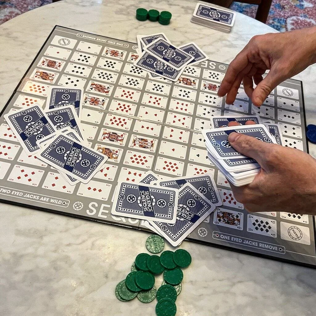 A game of Sequence in progress on a table showing cards, chips, and a game board with a grid of playing card images.