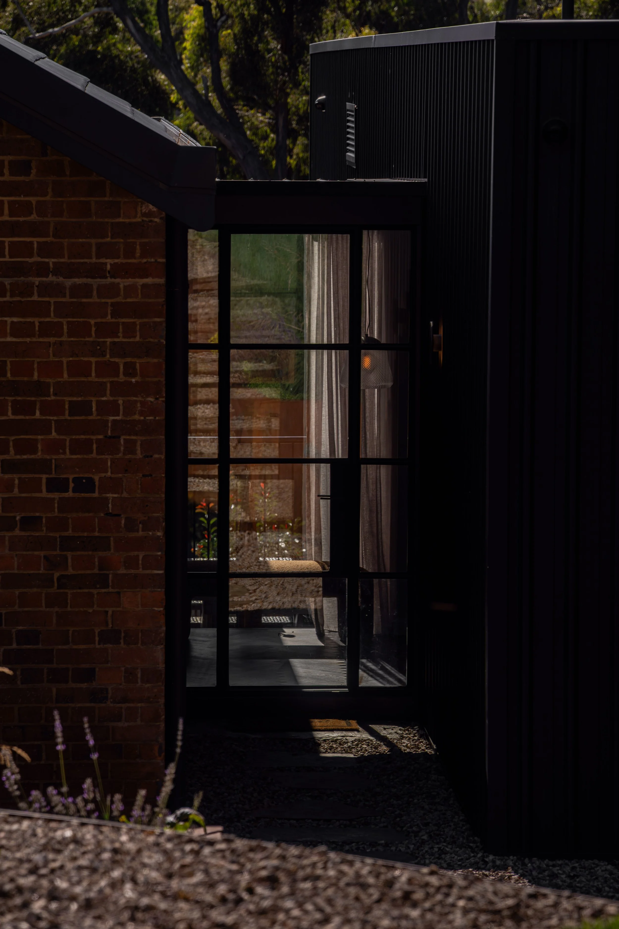 A view through a glass door with black framing, showing an indoor space with a chair, curtains, and a glimpse of greenery outside, taken during daytime.