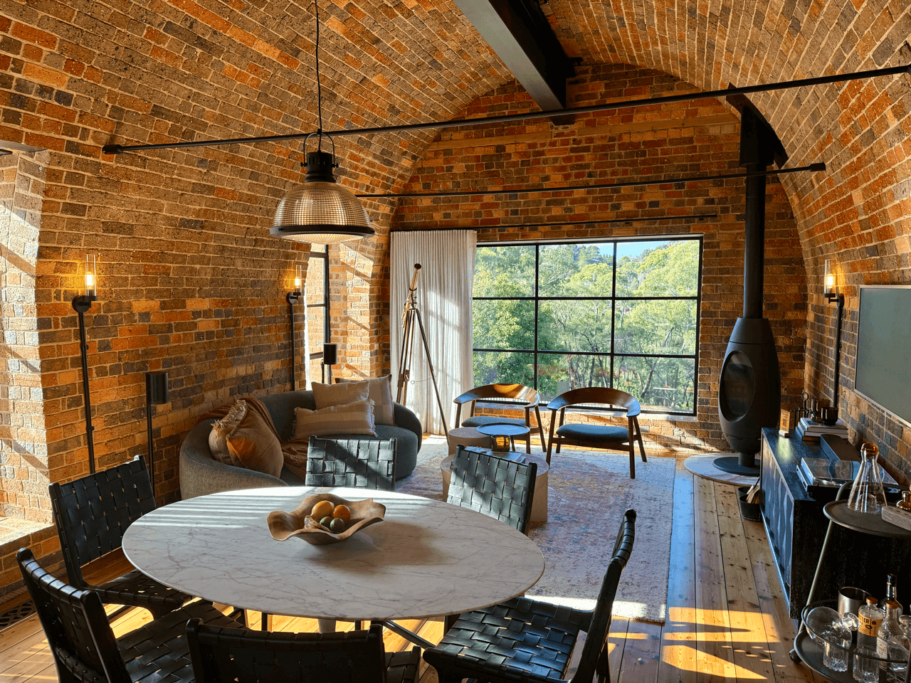 Living room with exposed brick walls, large window with view of trees, dining table with black chairs, gray sofa with pillows, two armchairs near the window, and a modern black fireplace.