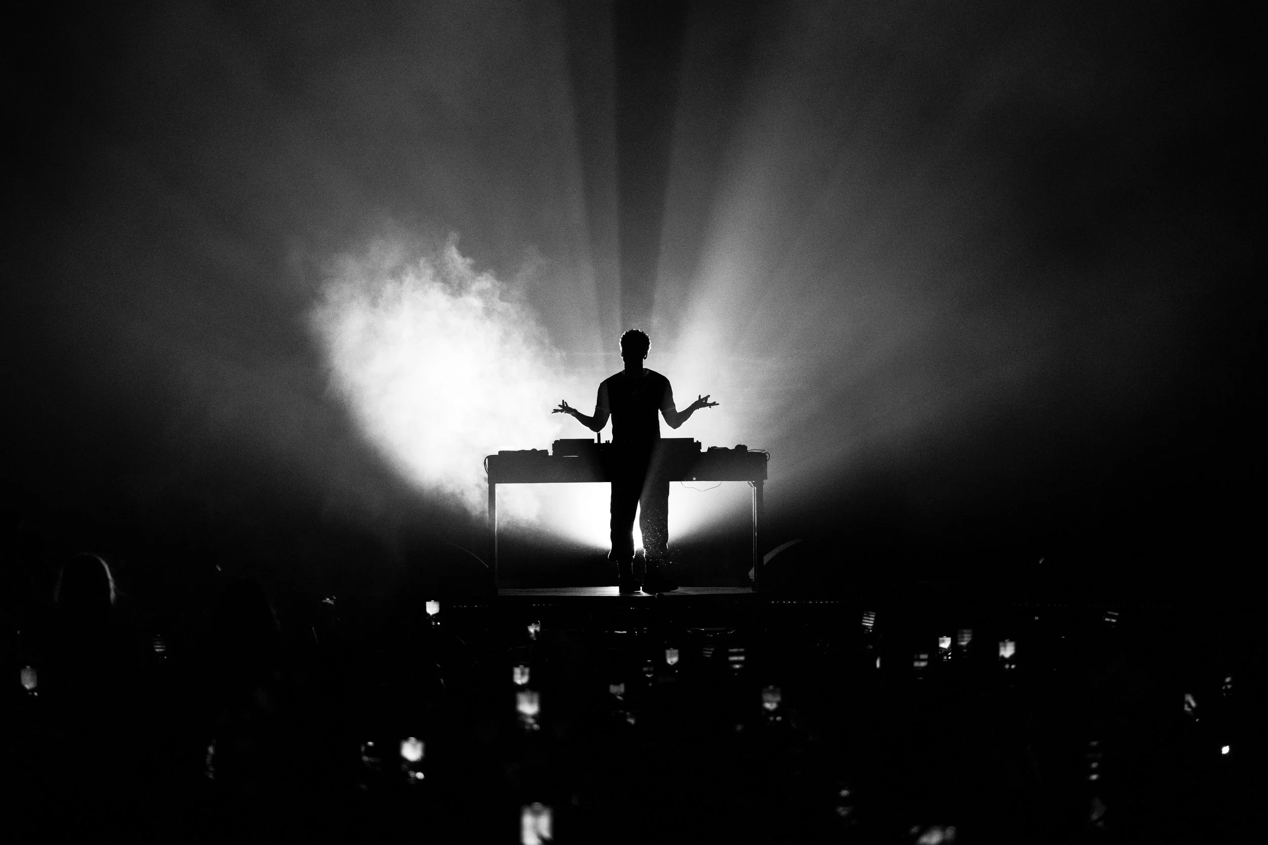 Silhouette of a person standing behind a DJ console on stage, illuminated by backlighting, with audience members holding up phones in the foreground.