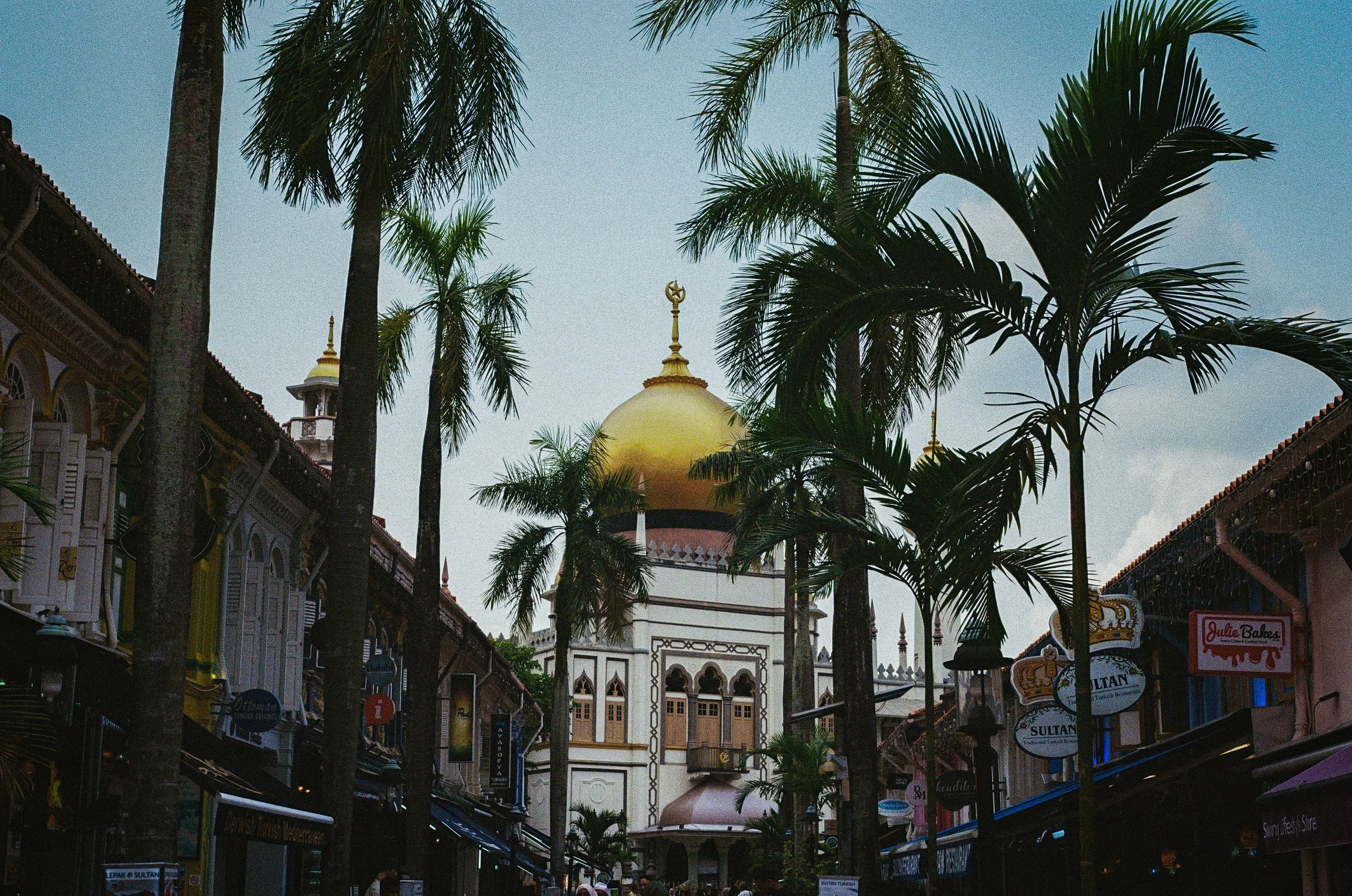 A busy street with palm trees lining both sides, leading to a building with a large golden dome, possibly a mosque, in the distance under a blue sky.
