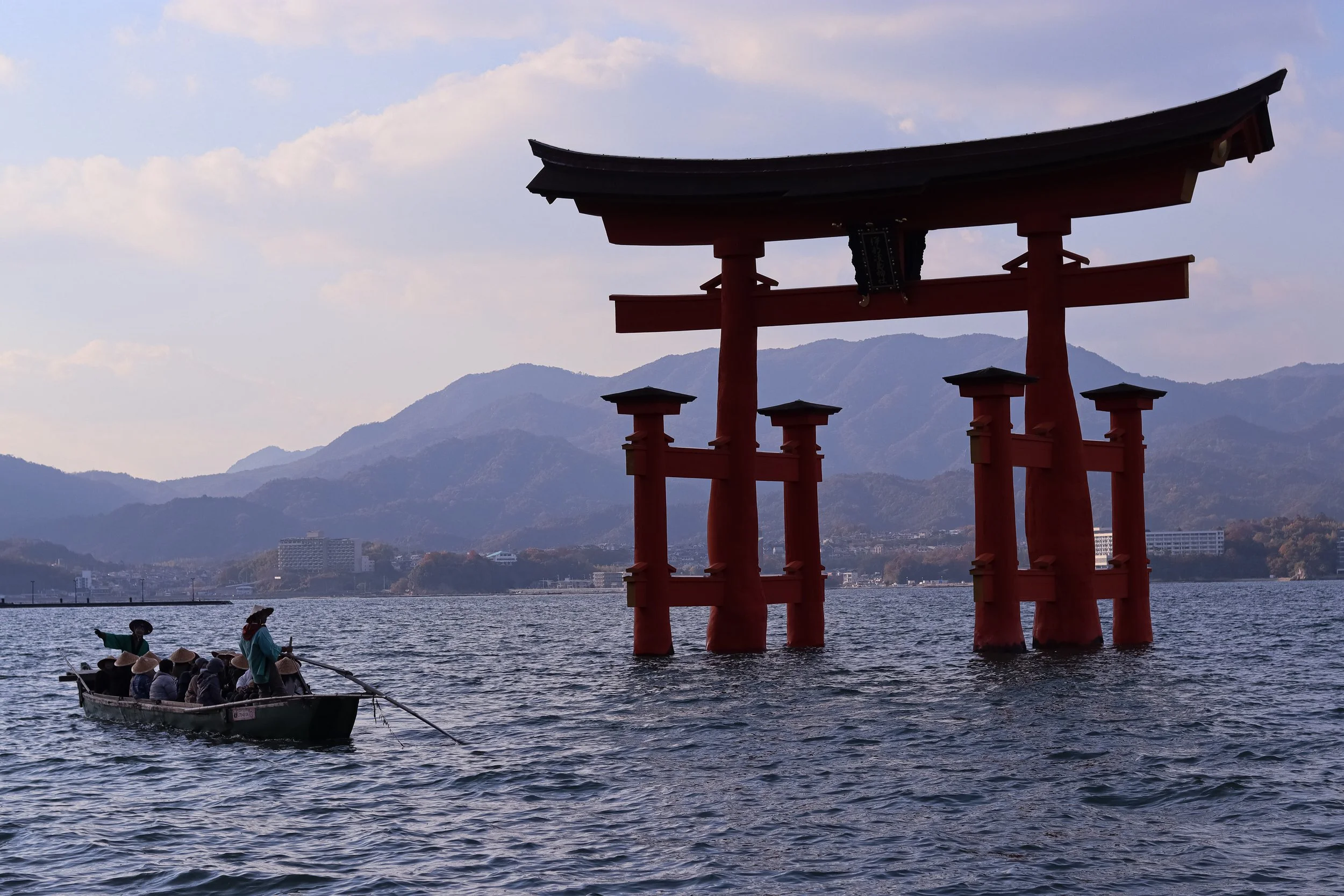 A floating red torii gate in a lake with mountains and a cloudy sky in the background, and a boat with people wearing traditional straw hats nearby.