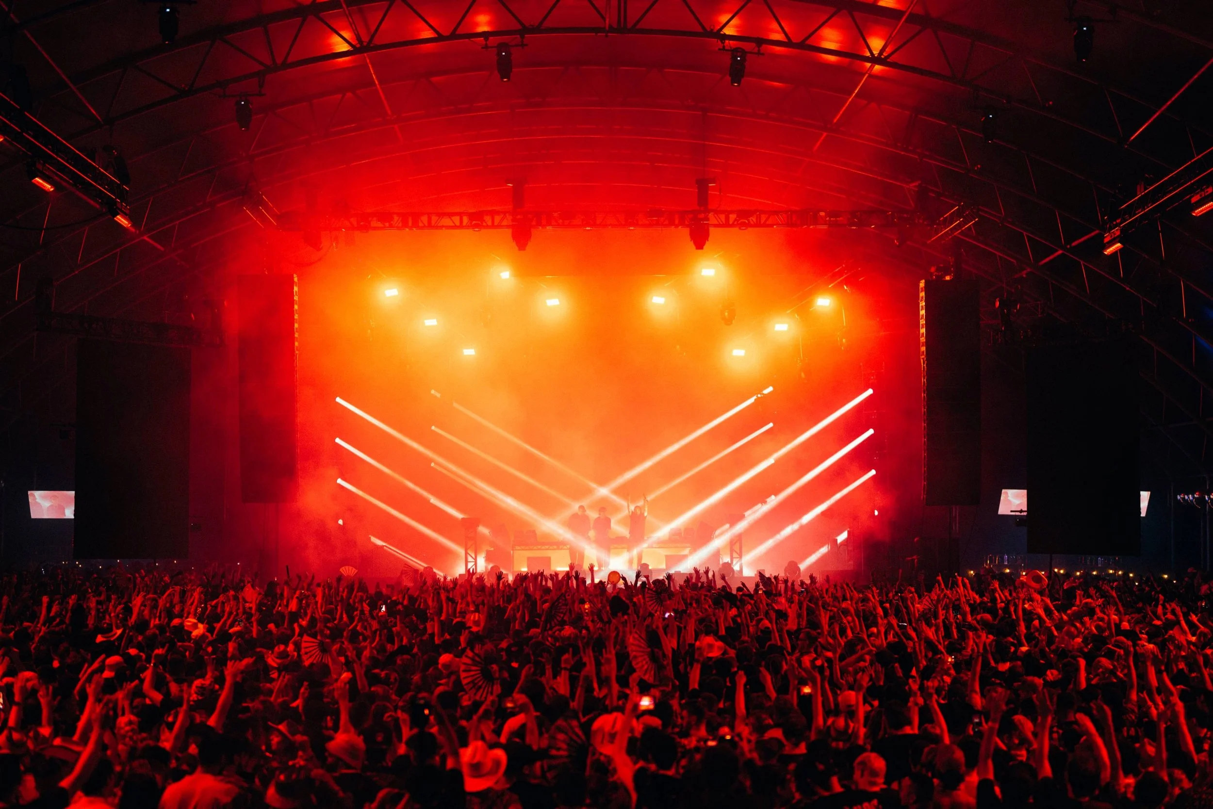 A concert or music festival with a large crowd in front of a brightly lit stage. The stage is illuminated with orange and yellow lights, with performers and dancers visible center stage. The audience has their hands raised, enjoying the live music performance.