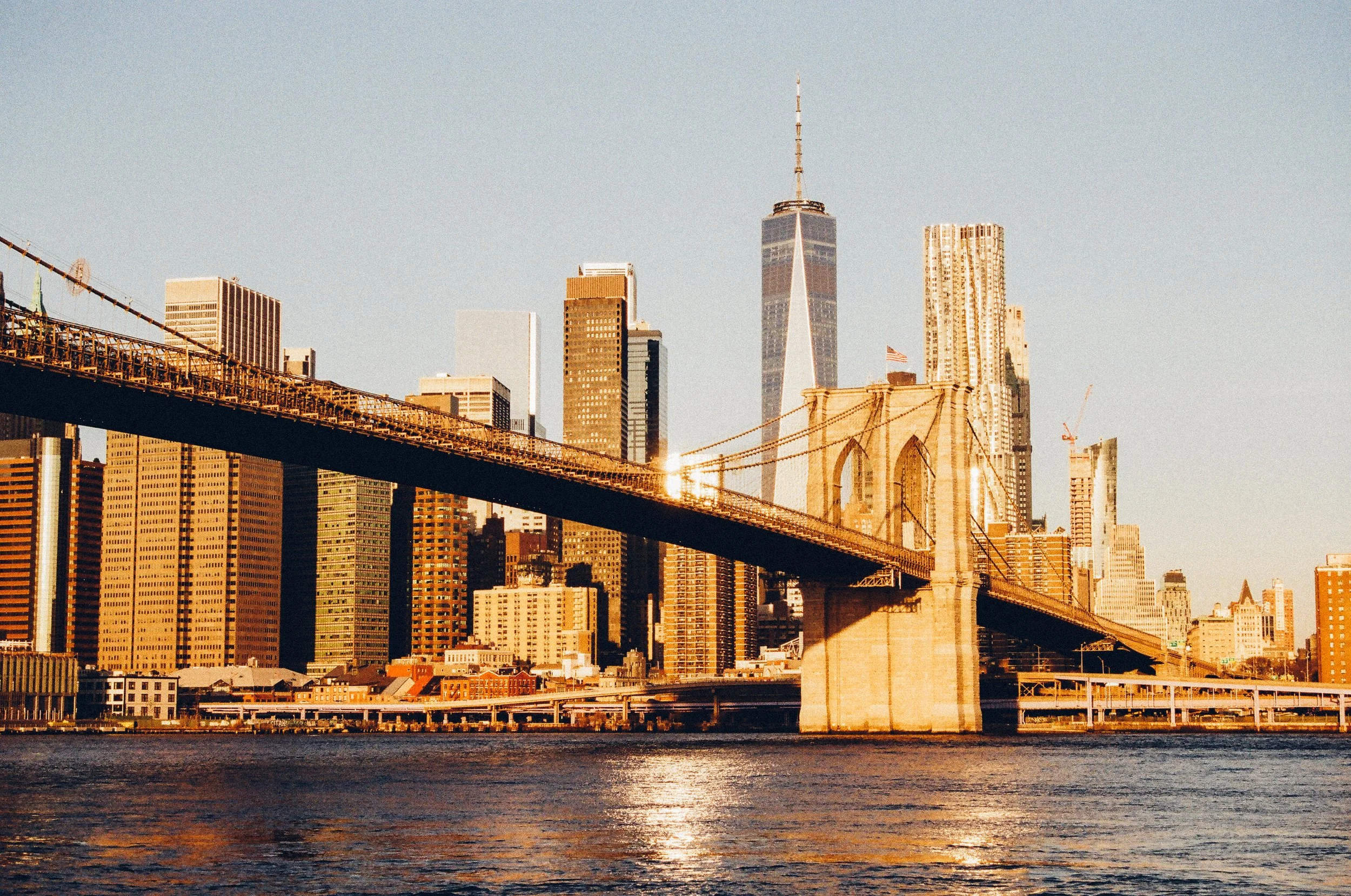 Sunlit view of the Brooklyn Bridge with New York City skyline in the background, including One World Trade Center, during sunset.