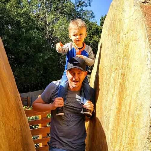 A young boy sitting on an adult's shoulders, smiling and waving, outdoors between two large tree trunks.