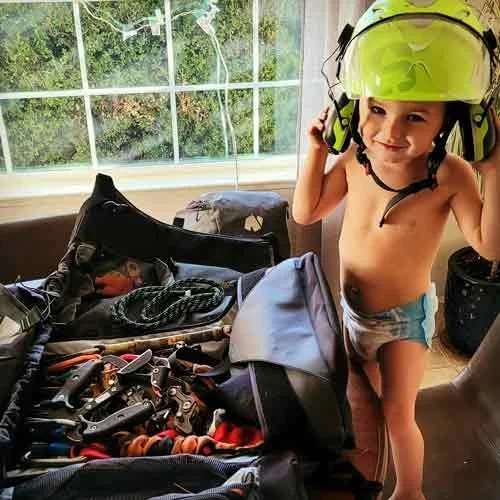 A young child with no shirt and shorts, putting on a large yellow safety helmet with headphones, standing next to an open suitcase filled with clothes and belongings, in a room with a large window showing trees outside.