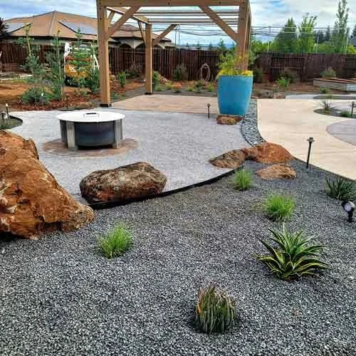 A modern backyard landscape featuring large rocks, small green plants, and gravel pathways under a wooden pergola.
