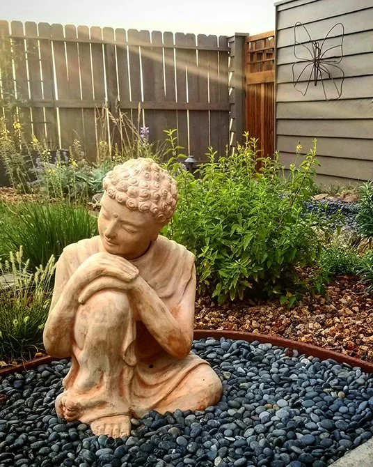 A garden statue of a young girl sitting with her arms resting on her knees and looking down, surrounded by plants and black rocks, with a wooden fence and house siding in the background.