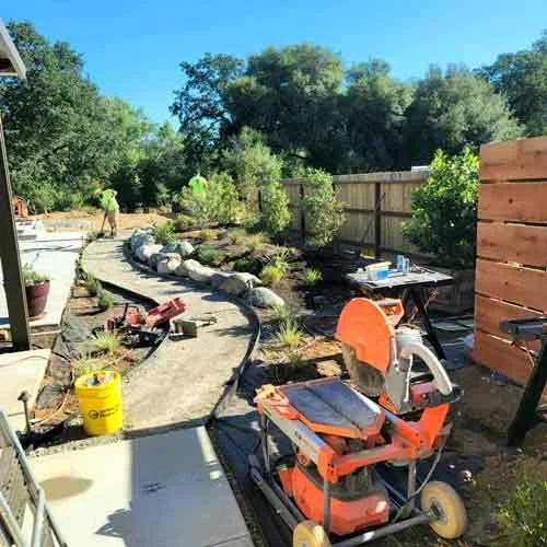 A backyard garden under construction with a curved stone pathway, construction tools, and workers in the background. There are trees, a wooden fence, and construction materials around.
