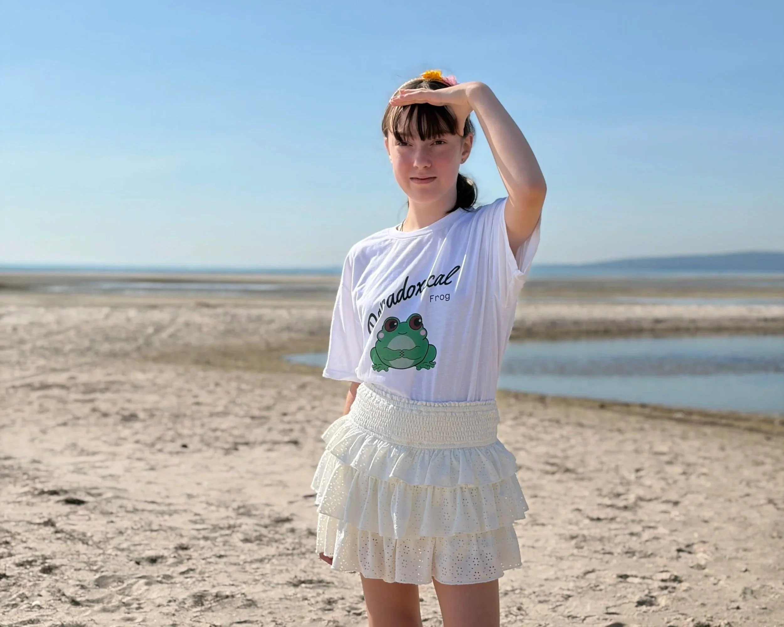 A young girl with dark hair and bangs standing on a beach, wearing a white T-shirt with a cartoon frog and a white ruffled skirt, shielding her eyes from the sun with her hand.