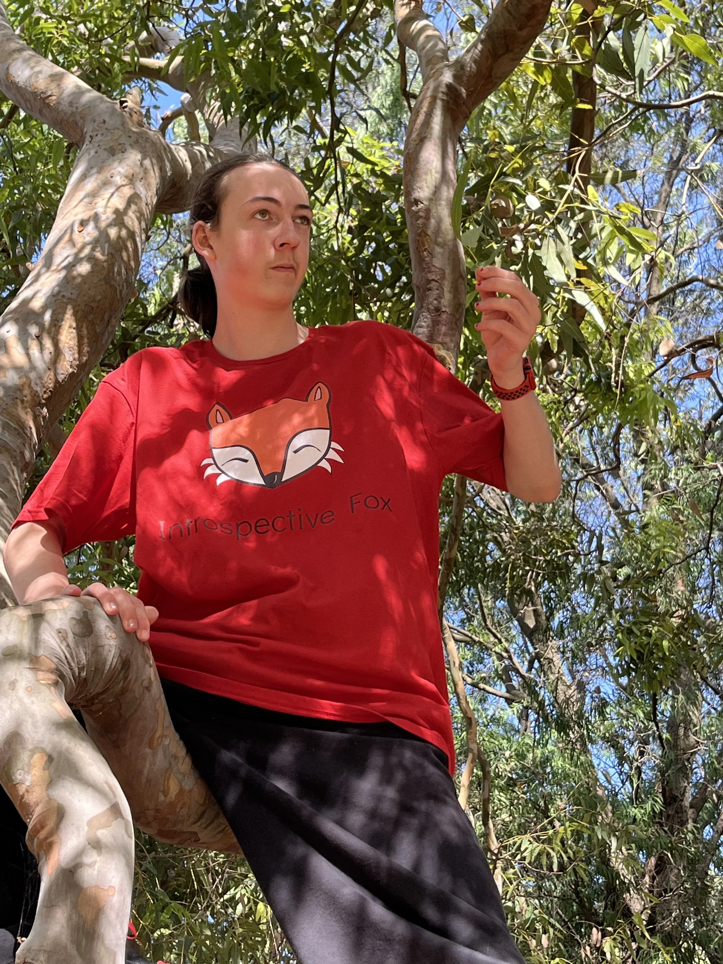 A woman with dark hair in a ponytail is sitting in a tree, surrounded by green leaves and branches. She is wearing a red T-shirt with a fox graphic and the words 'Intraspective Fox' on it, and black pants. She is looking to the left with a neutral ex