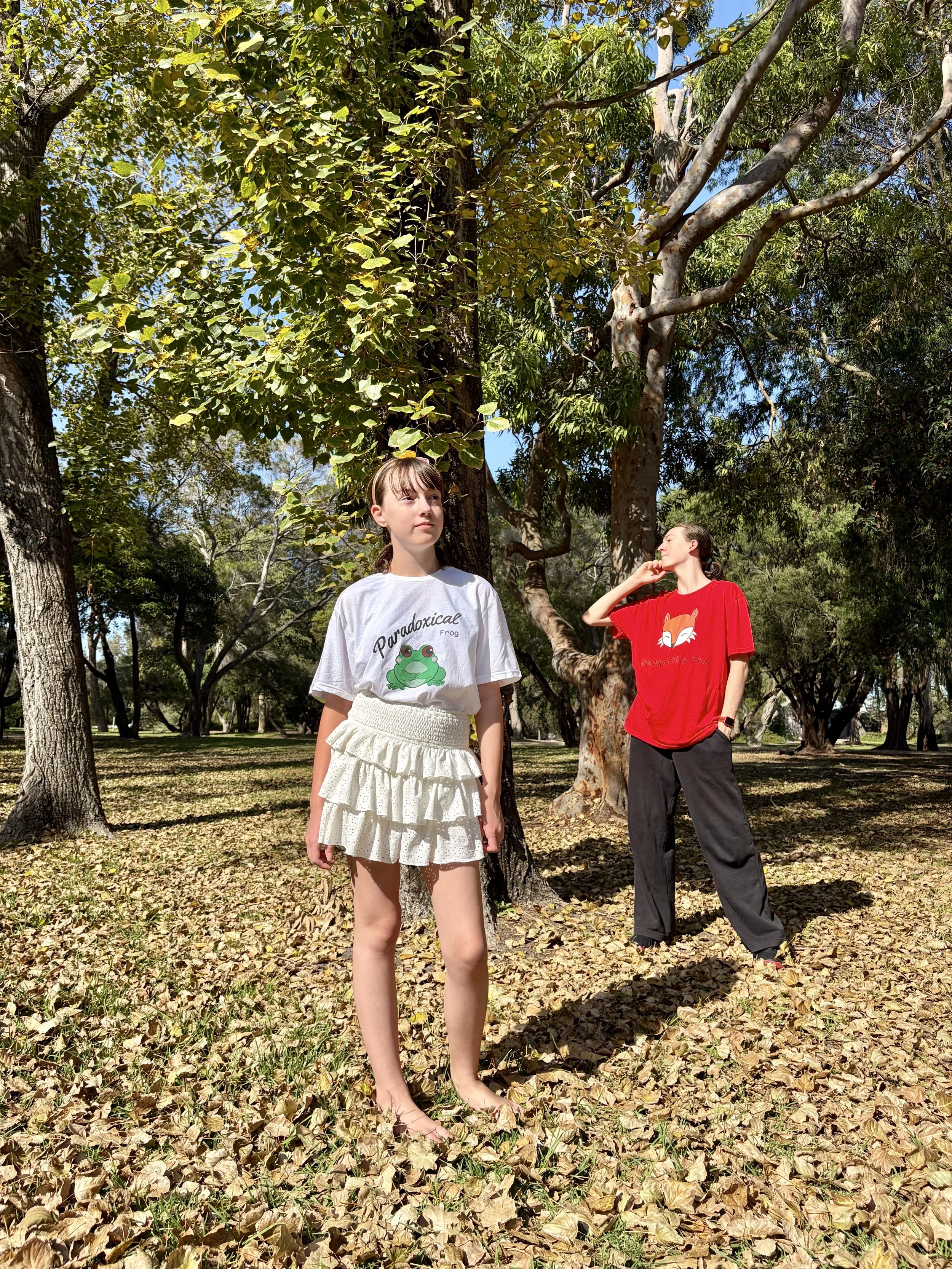 Two young women standing outdoors in a park with trees and fallen leaves. One woman in a white T-shirt and skirt is barefoot, looking off to the side. The other woman in a red T-shirt and black pants is scratching her face, looking upward.