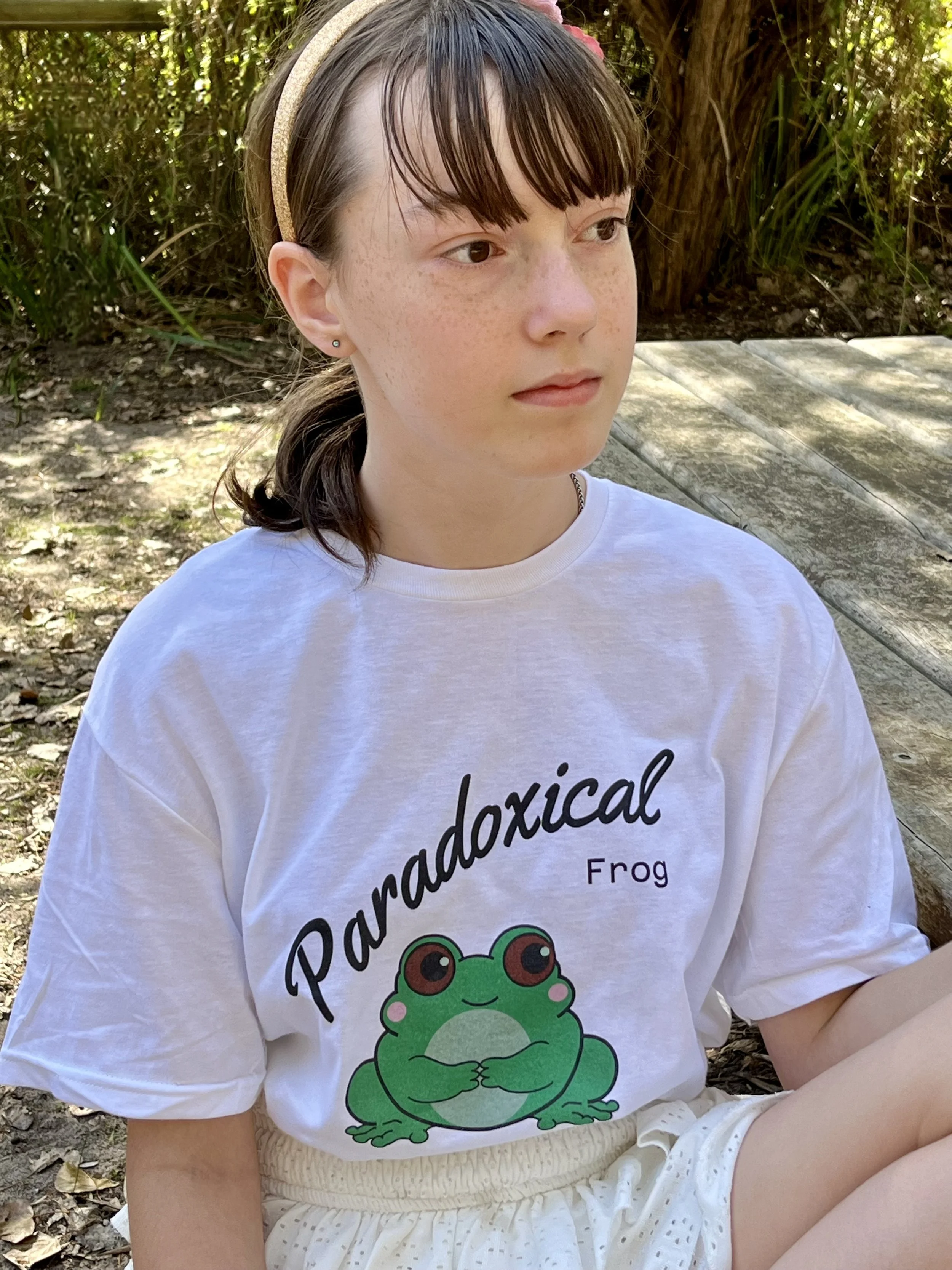 Close-up of a young girl with brown hair, wearing a headband and a T-shirt with a frog graphic and the words 'Paradoxical Frog'. She is outdoors, surrounded by trees and wooden benches.