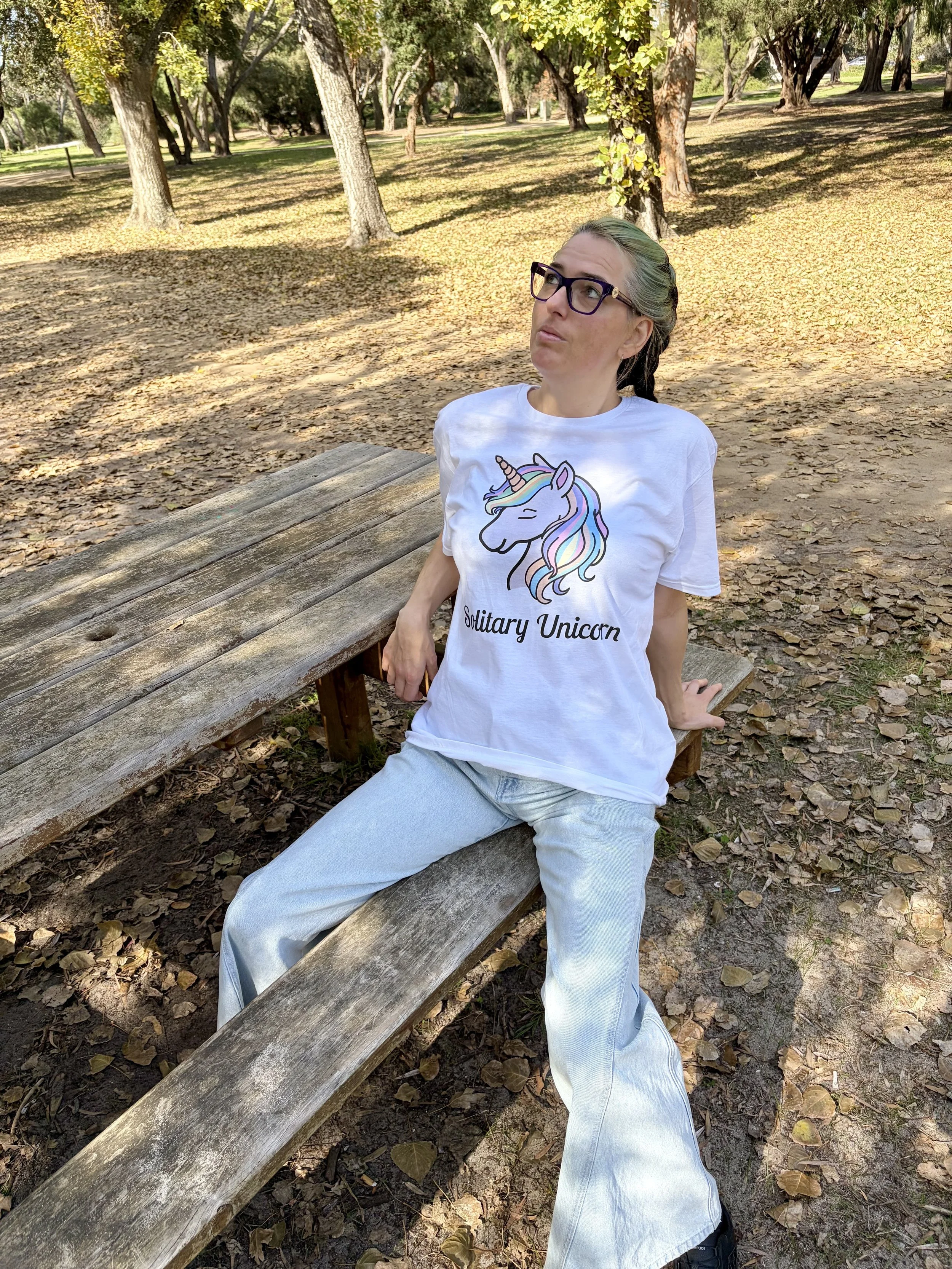 A woman with glasses and a tied-back hairstyle is sitting on a wooden park bench in a wooded area with trees and fallen leaves, wearing a white T-shirt with a colorful unicorn graphic and the words "Solitary Unicorn," and light-colored jeans.