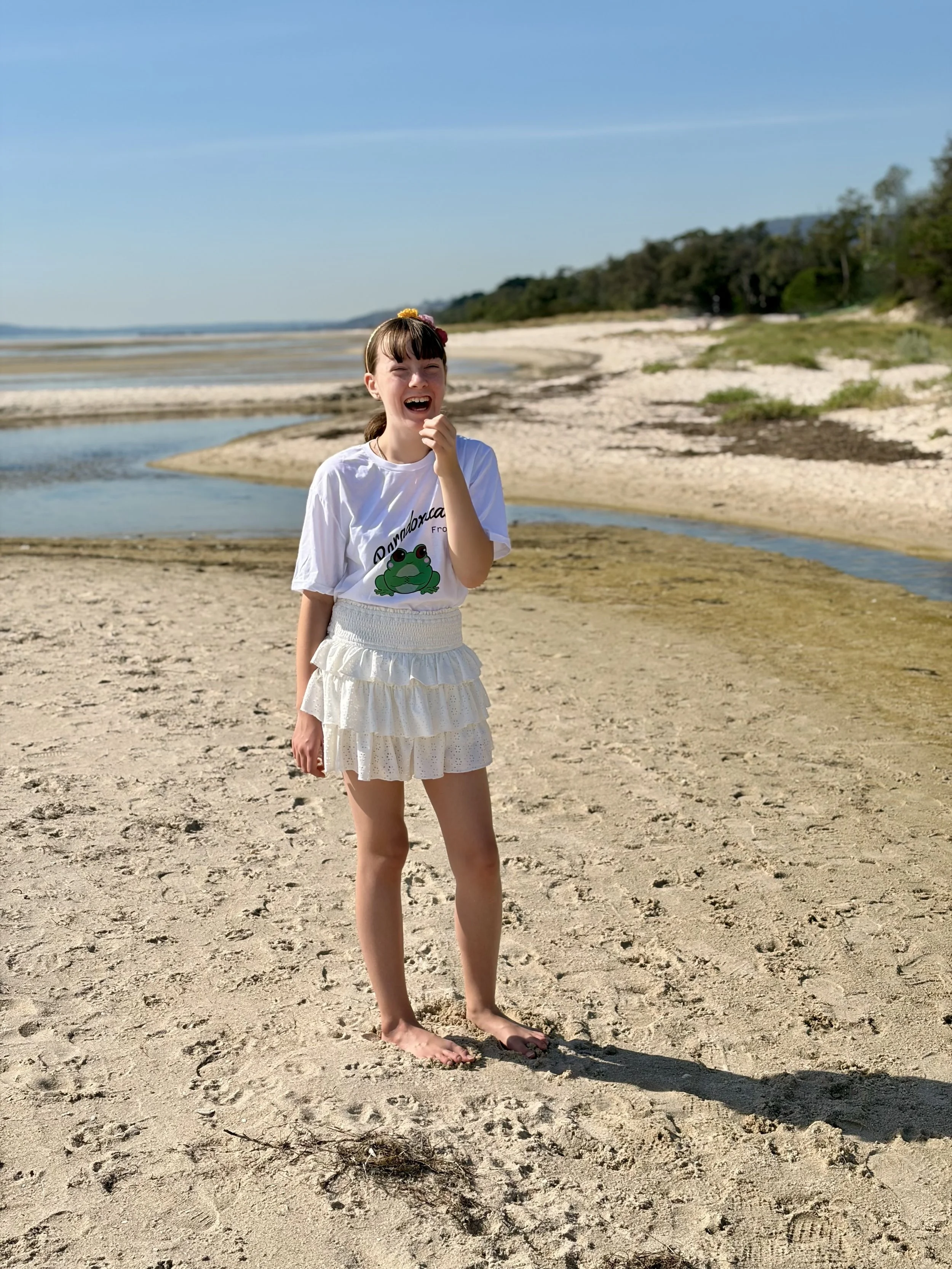 A young girl standing on a sandy beach near water, laughing and wearing a white T-shirt with a frog graphic and white ruffled skirt.