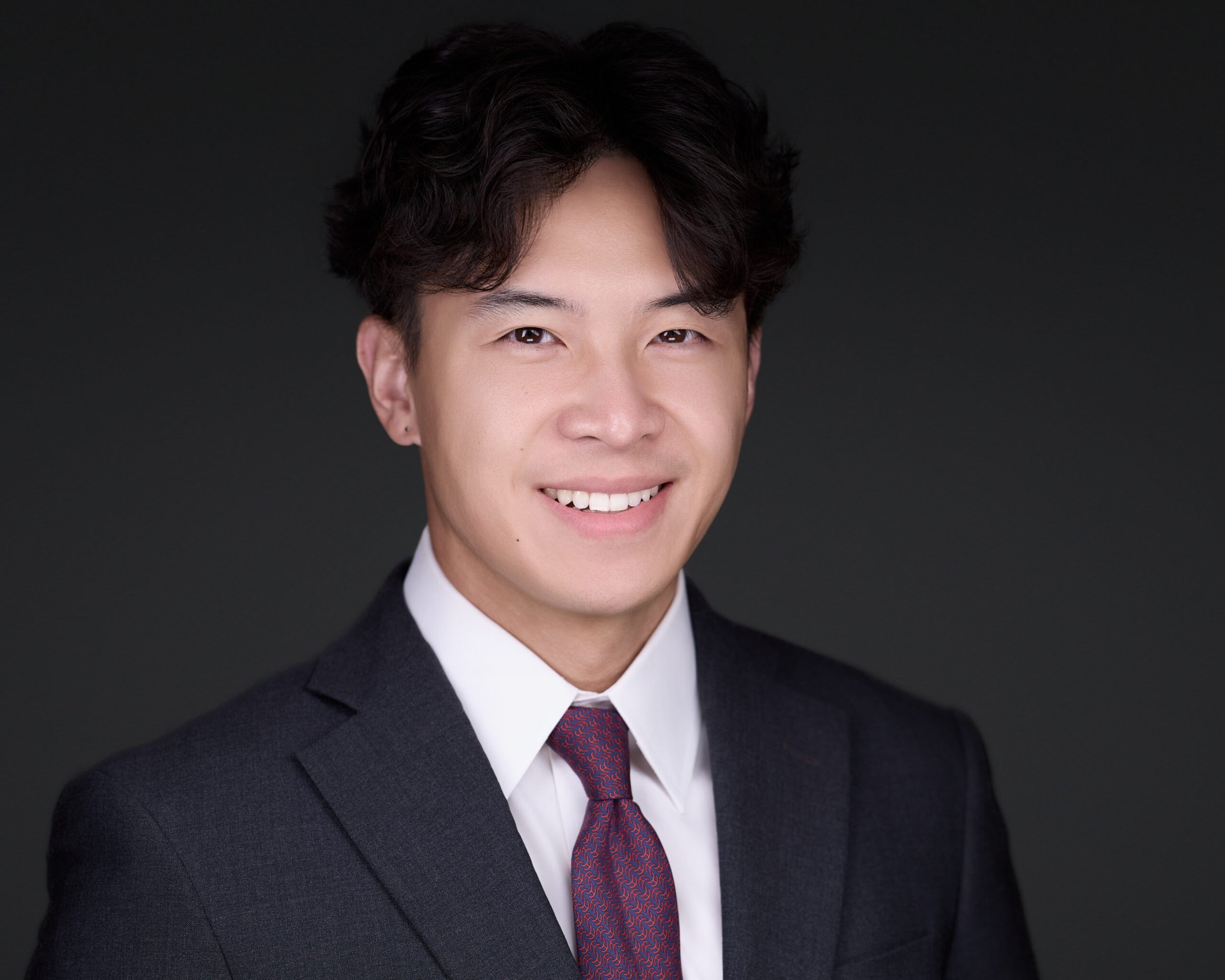 Professional headshot of a smiling young Asian man in a dark suit, white shirt, and patterned tie against a dark background.