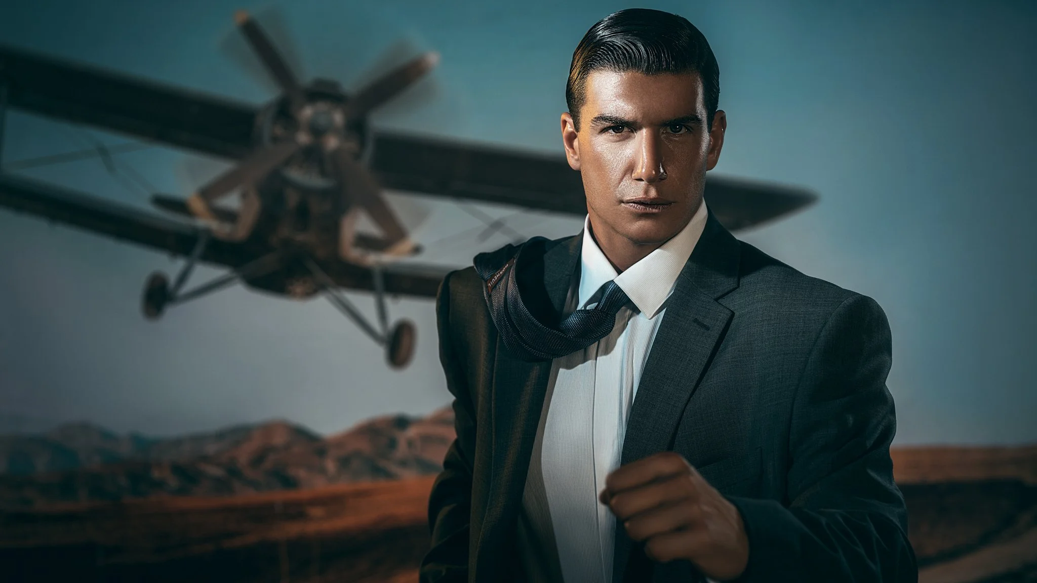 A man in a suit with a scarf around his neck standing in front of a model airplane in a desert landscape.