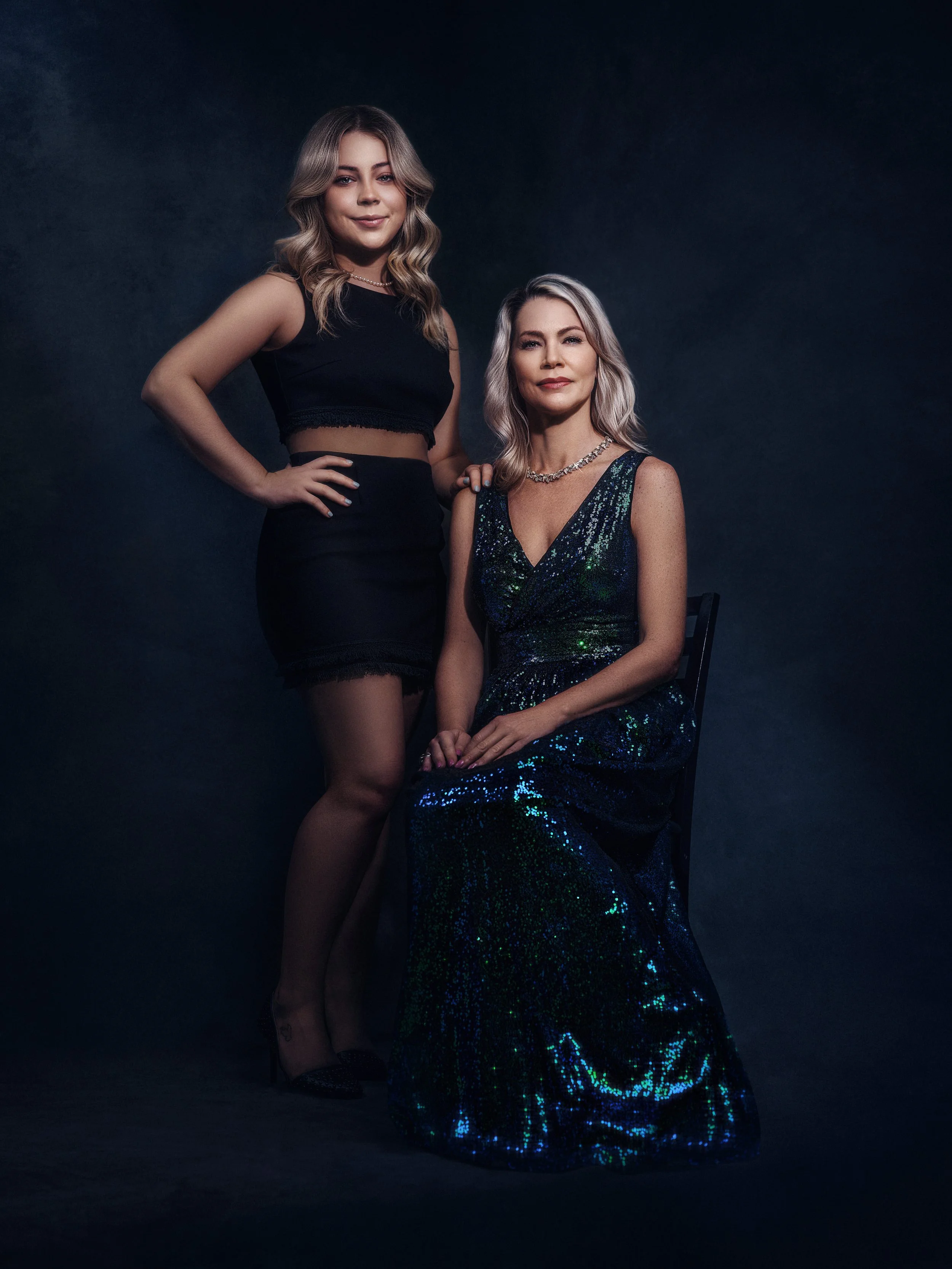 Two women in elegant evening wear, one standing and one seated on a chair, posing against a dark studio background.