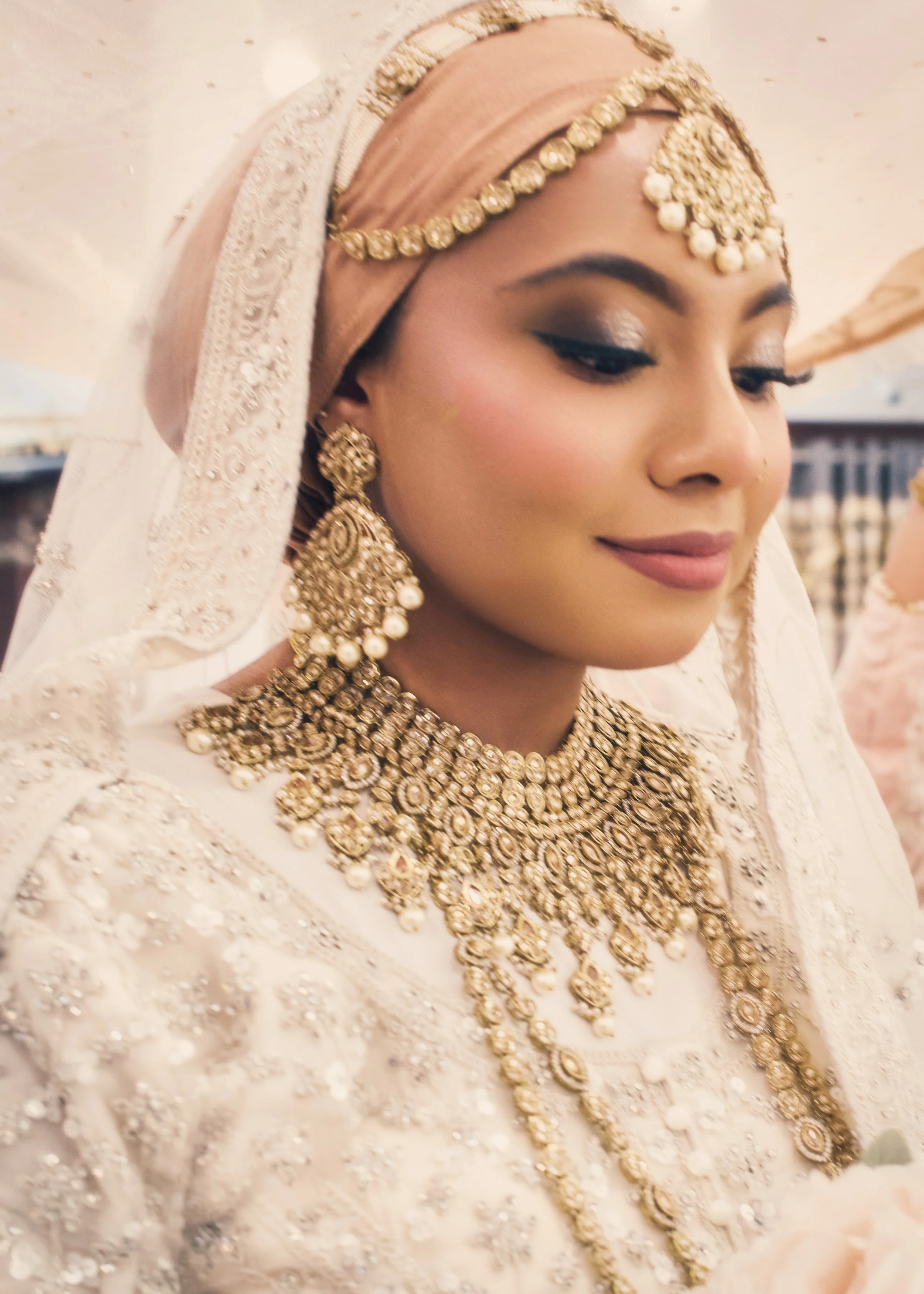 A woman in traditional wedding attire with elaborate gold jewelry, including a layered necklace, large earrings, and a decorative headpiece, smiling softly.