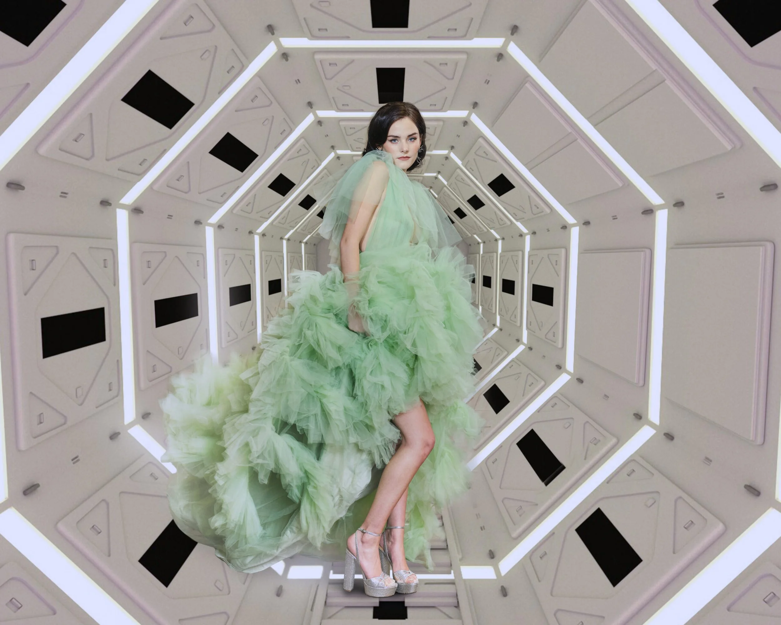 A woman in a flowing, ruffled, light green dress and silver high heels stands inside a futuristic, geometric tunnel with glowing white edges and panels with black rectangles.