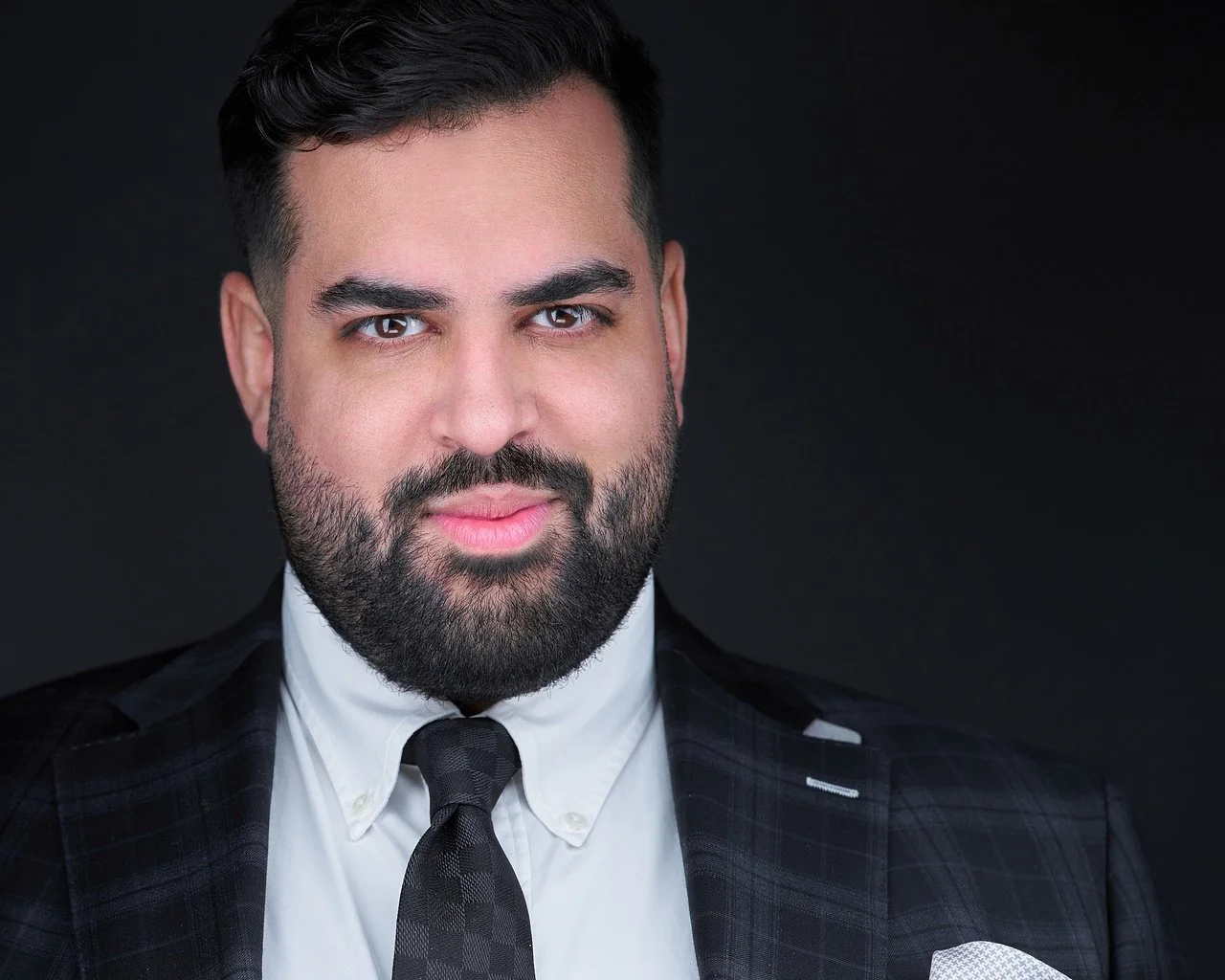 A portrait of a man with dark hair, a beard, wearing a dark plaid suit, white shirt, and a black tie, against a black background.