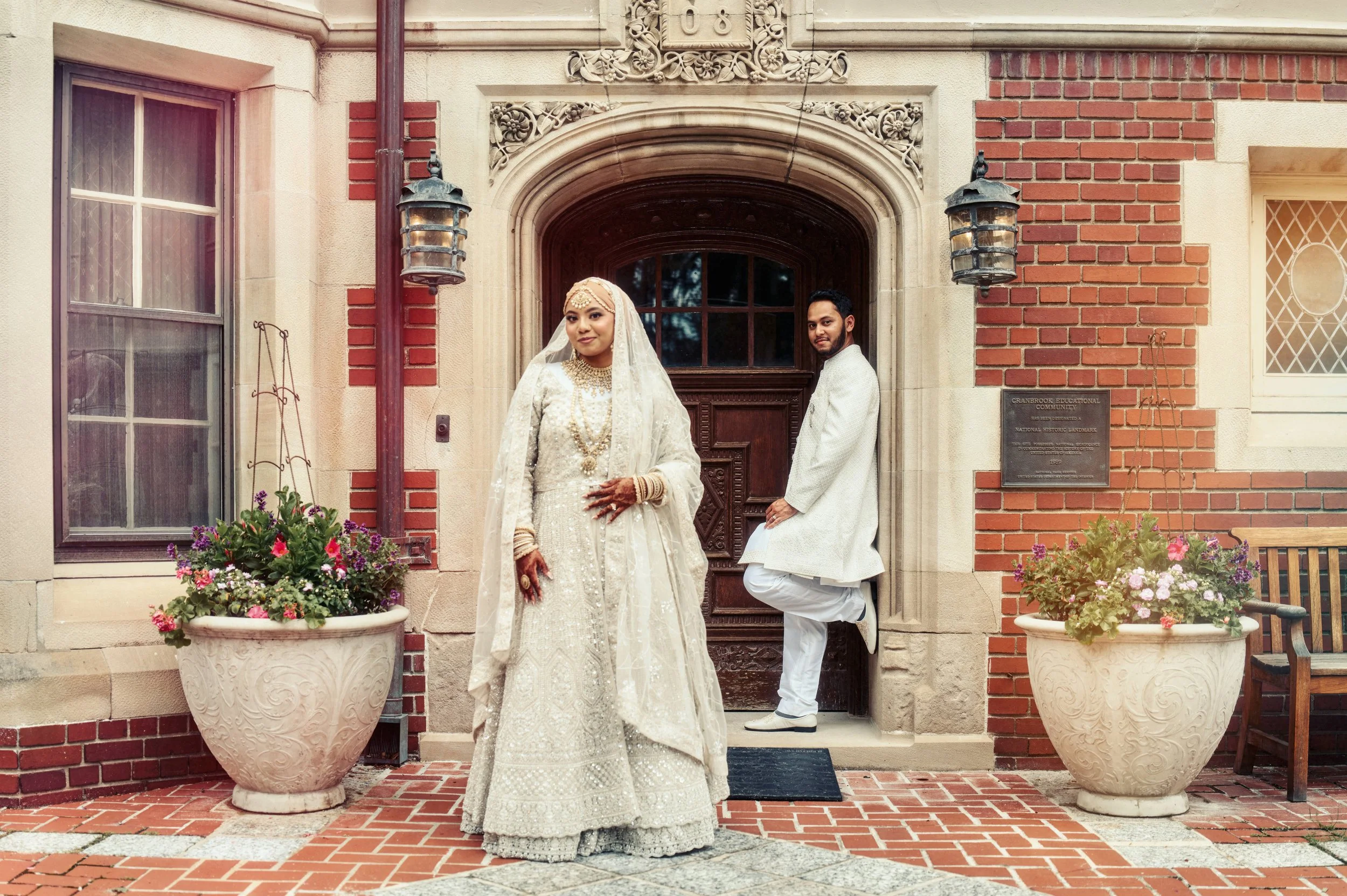 A couple in traditional Indian wedding attire standing outside a historic building with brick and stone facade, floral arrangements in large white planters, and ornate lanterns.
