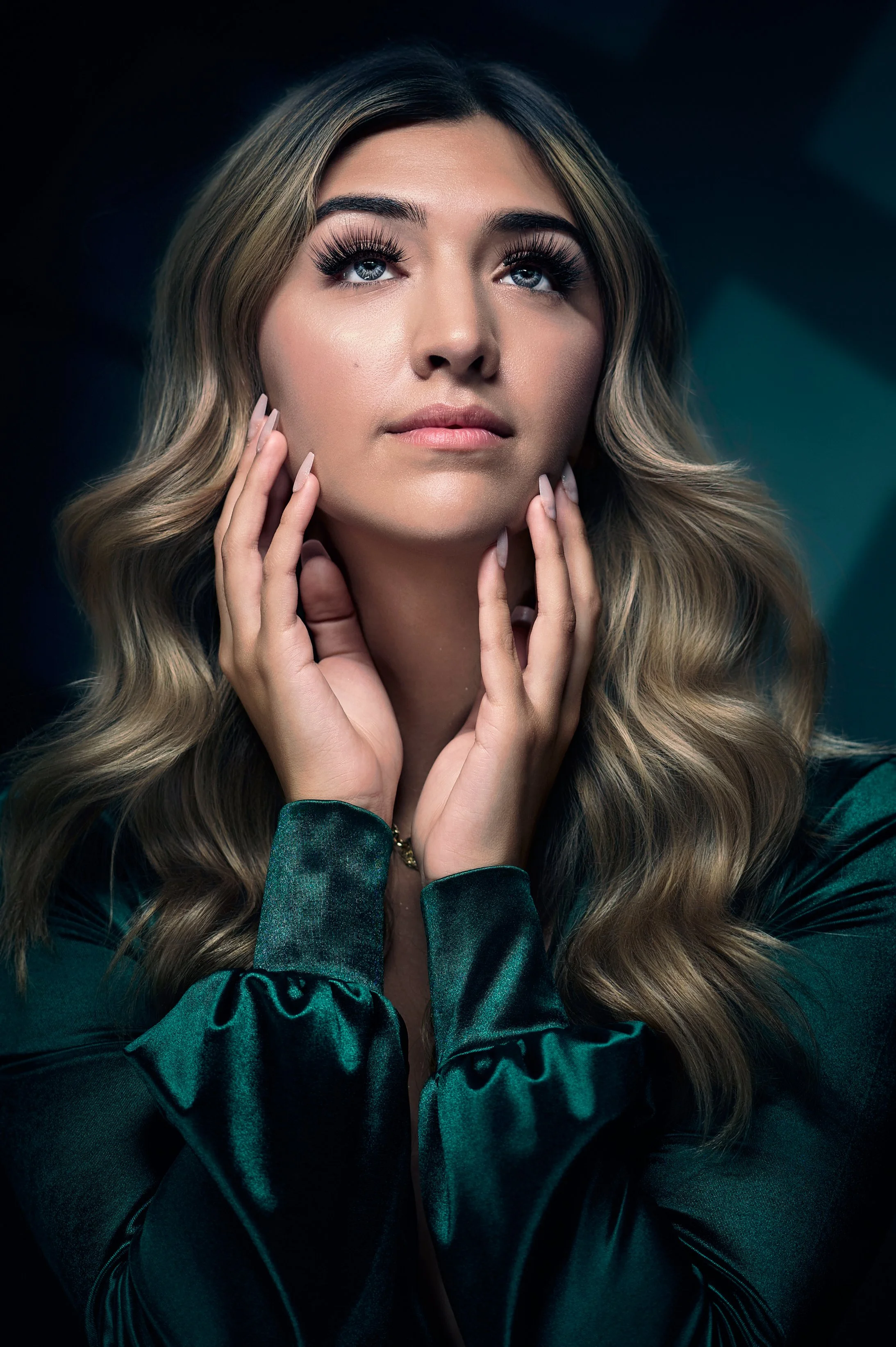 Portrait of a young woman with wavy blonde hair and blue eyes, wearing a dark green satin blouse, touching her face gently.