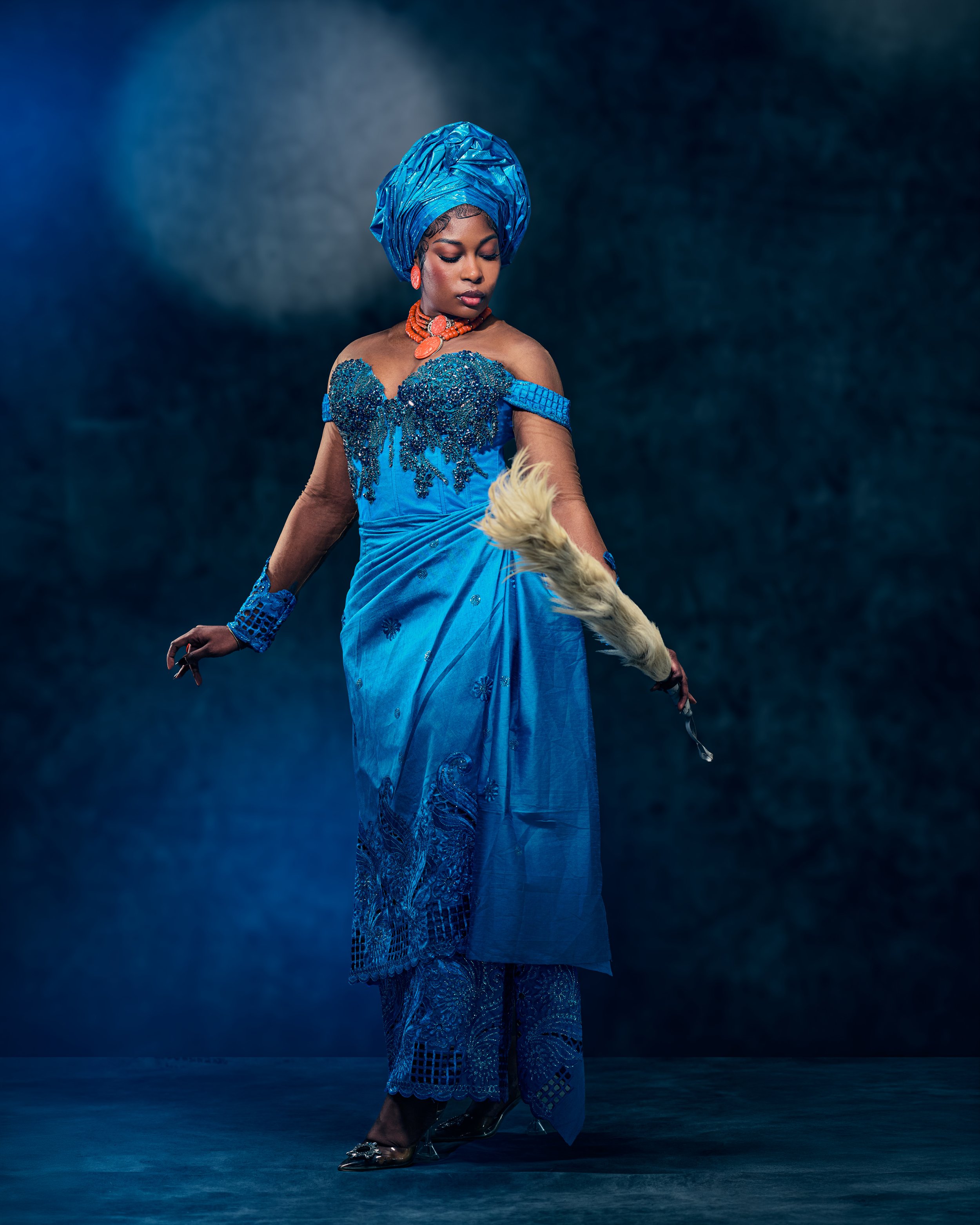 A woman in a blue traditional African dress and headwrap, holding a feather duster, standing against a dark background.