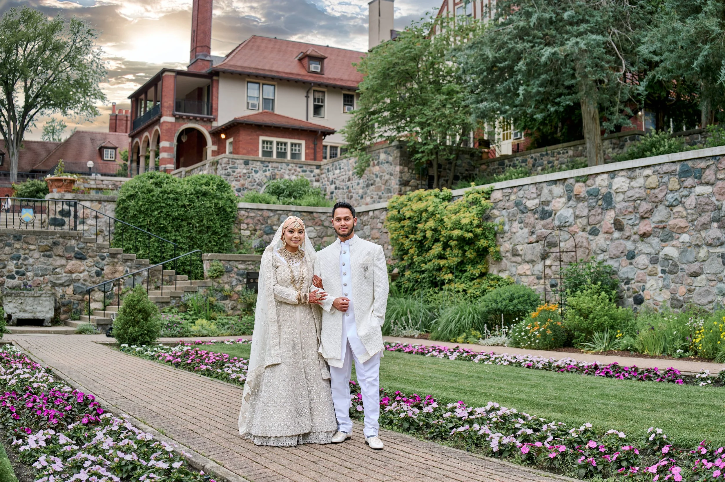 A bride and groom standing on a garden path outside a large house during sunset, with flowers and greenery surrounding them.