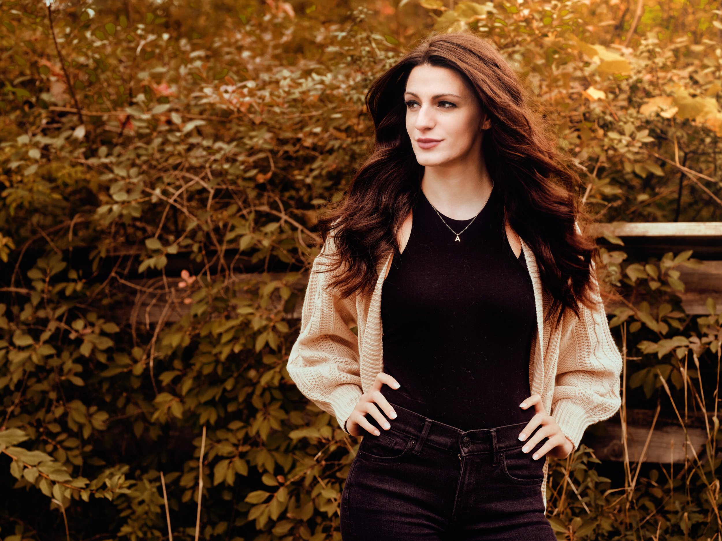 A woman with long, wavy brown hair stands outdoors with her hands on her hips, wearing a black shirt, a beige cardigan, and black pants, with autumn foliage in the background.