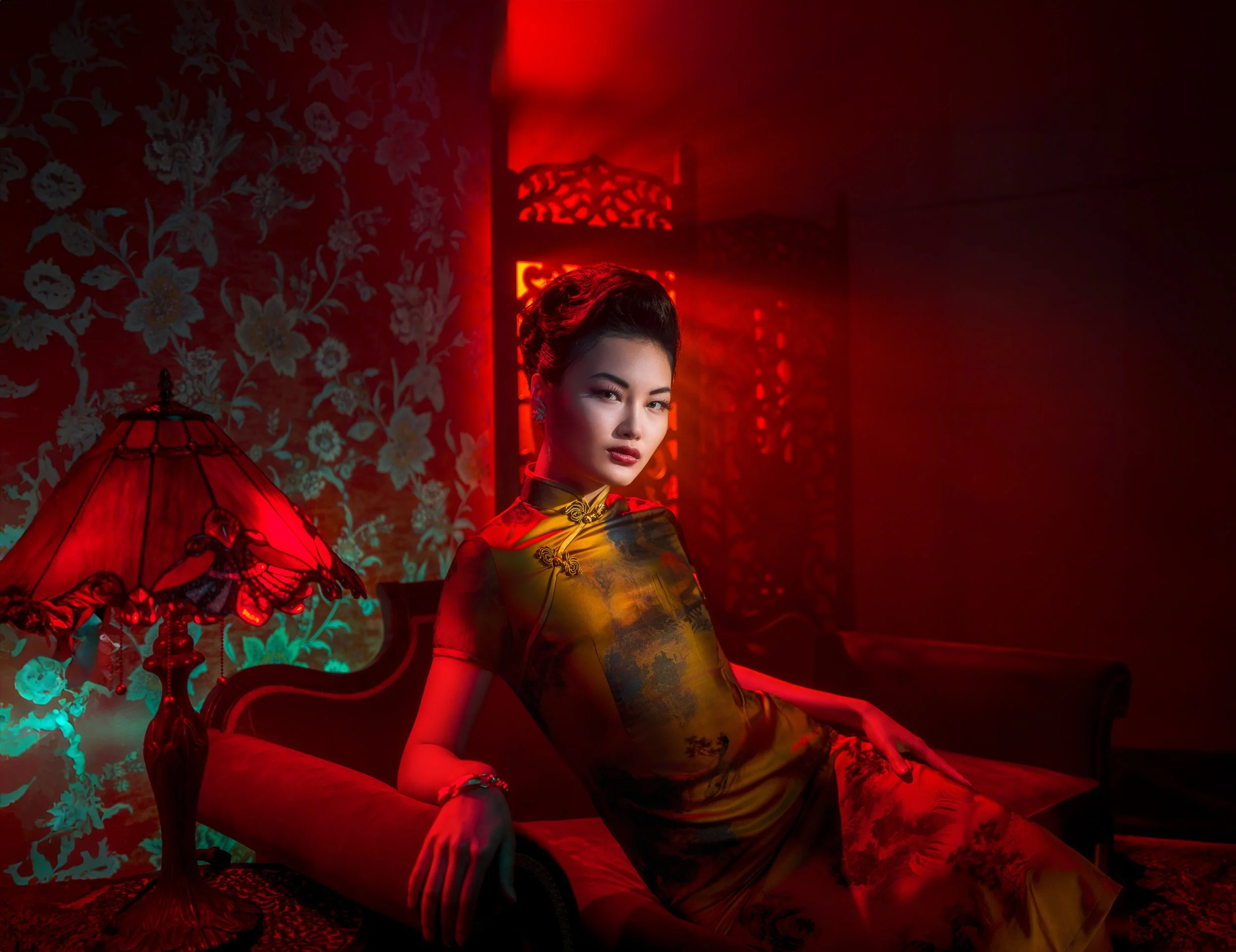 A woman in a traditional cheongsam dress sitting on an ornate sofa in a dimly lit room with red lighting and Asian decor, including a decorative screen and a stained-glass lamp.