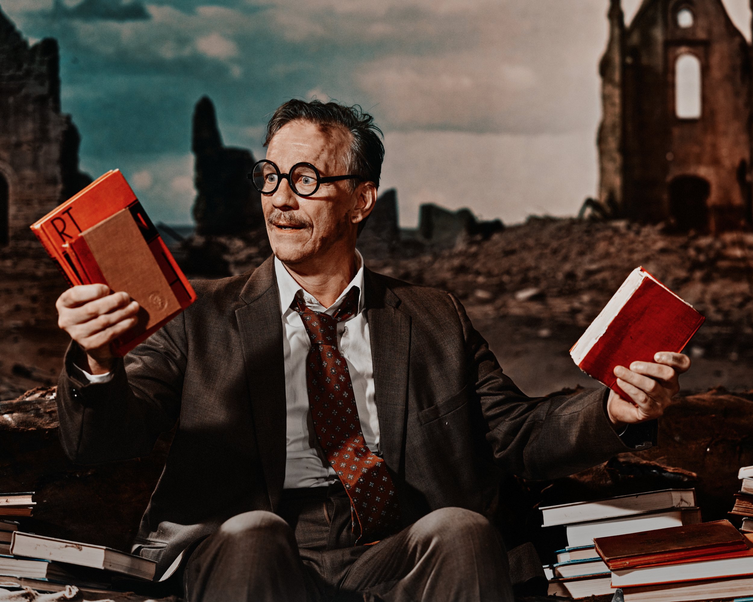 A man in a suit and glasses sitting amongst books on a desk, with a ruined building in the background.