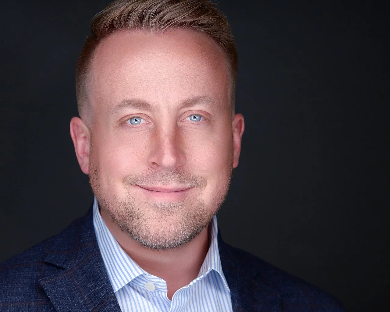 A professional headshot of a man with light skin, blue eyes, short blond hair, and a trimmed beard, wearing a striped collared shirt and a dark blazer, smiling against a dark background.
