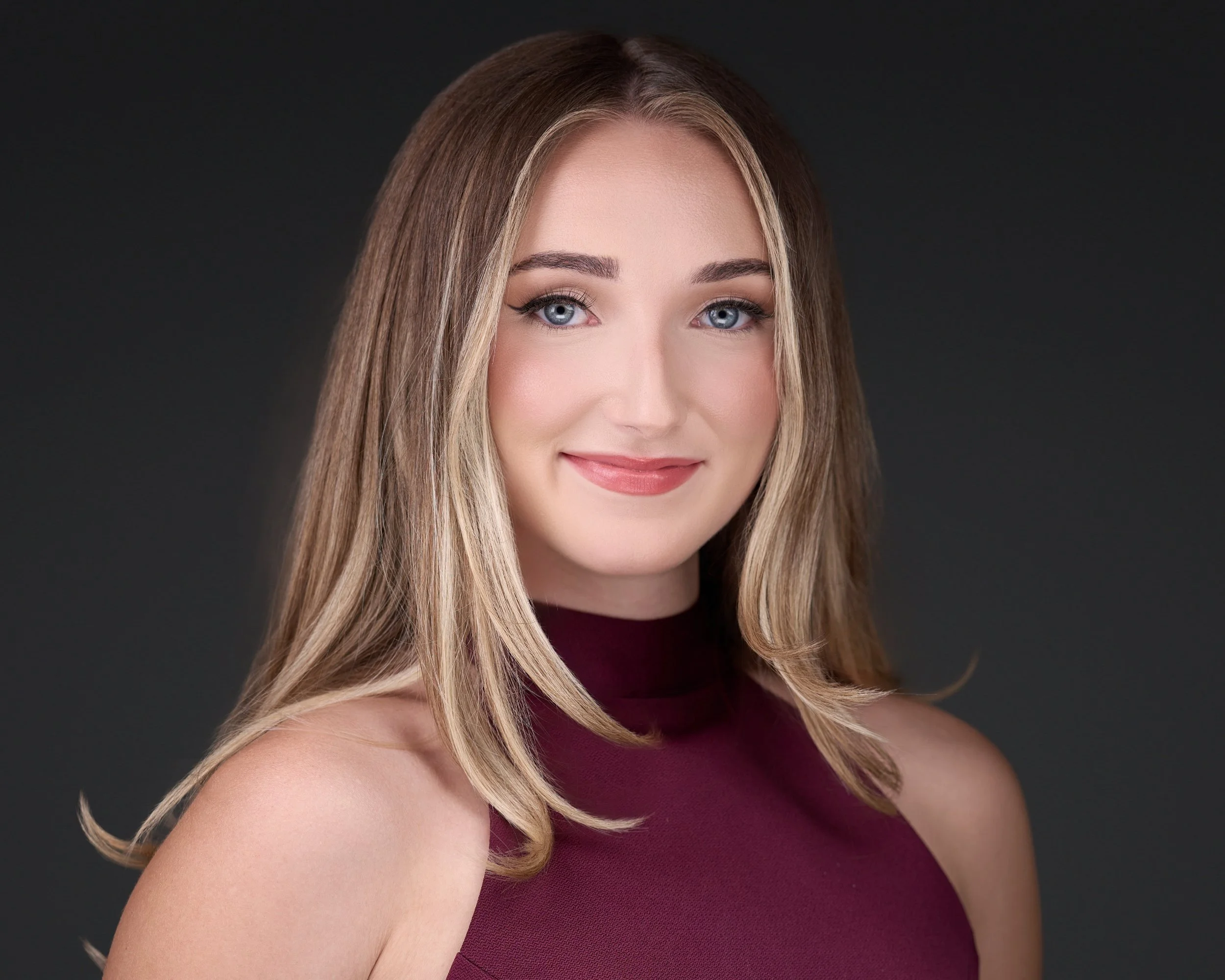 A woman with long, wavy blonde hair, blue eyes, and a warm smile, wearing a sleeveless maroon top against a dark background.
