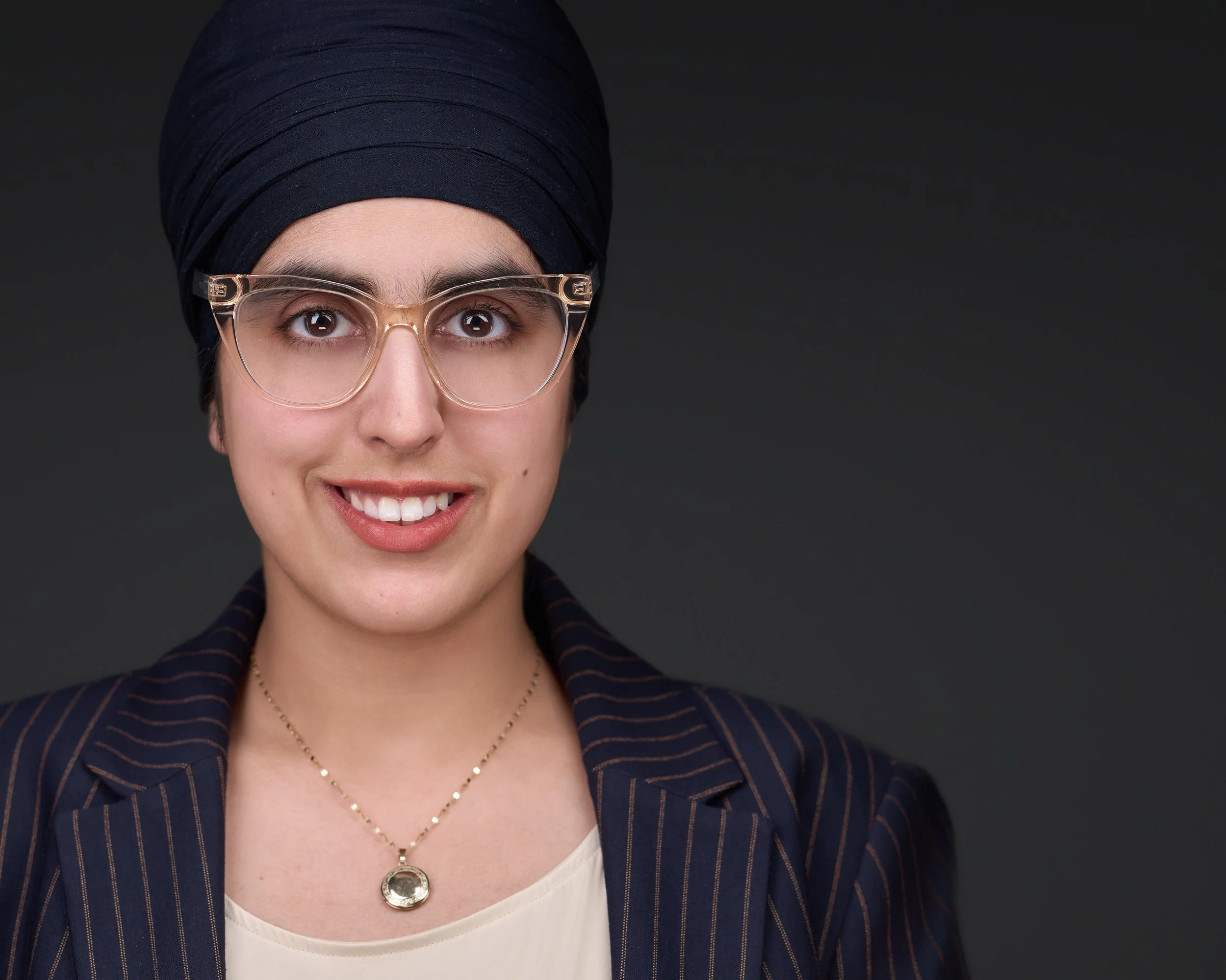 A woman with short dark hair wearing glasses, a black turban, a striped blazer, and a gold necklace, smiling against a dark background.