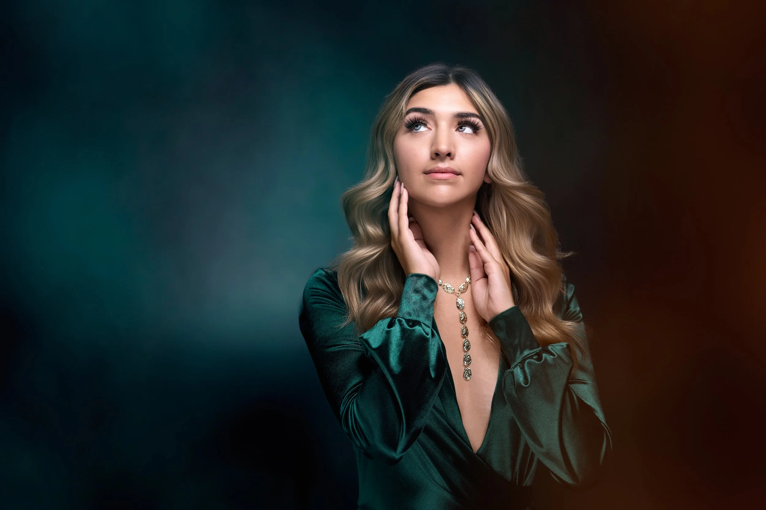 A young woman with long wavy blonde hair and light skin, wearing a satin green dress and a matching necklace, poses with her hands touching her neck and her head slightly tilted, against a dark, colorful gradient background.