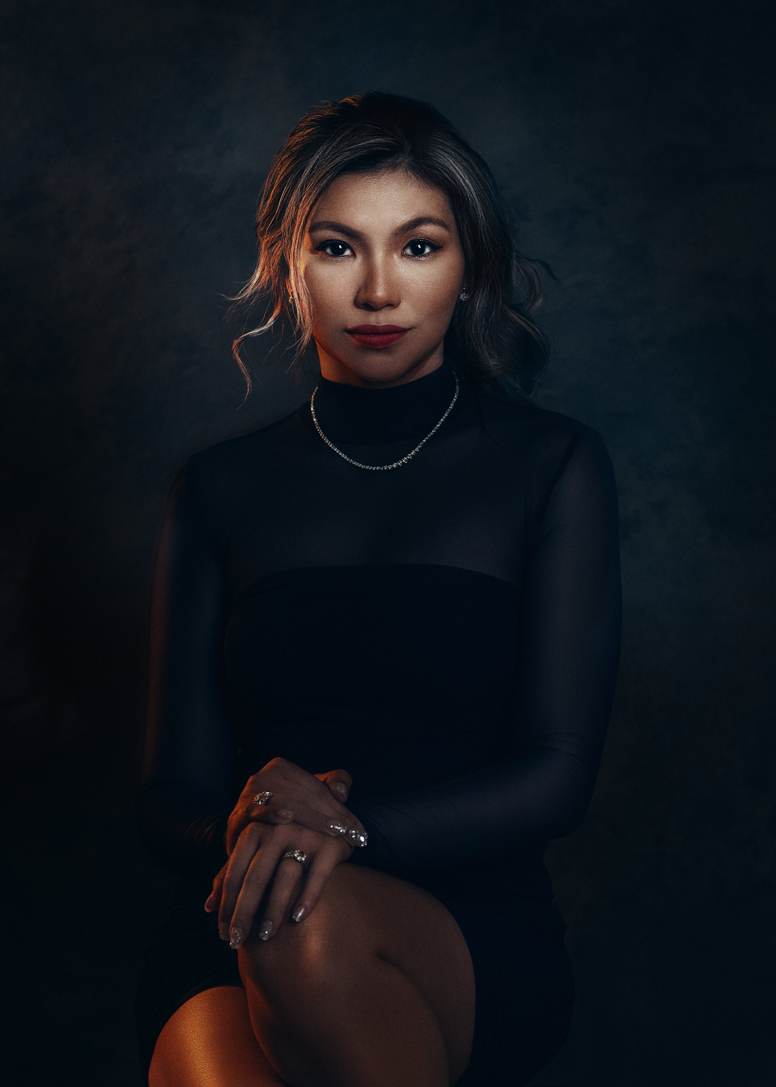 Portrait of a woman with dark hair wearing a black turtleneck dress, jewelry, and sitting against a dark background.