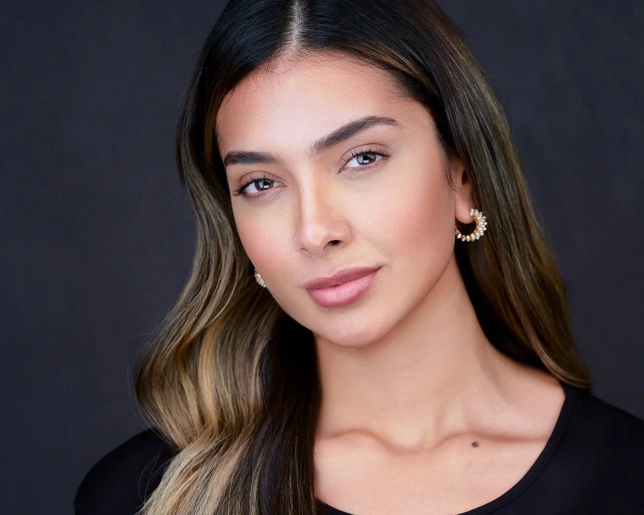 Close-up of a young woman with long wavy hair, wearing pearl earrings and a black top, looking confidently at the camera against a dark background.